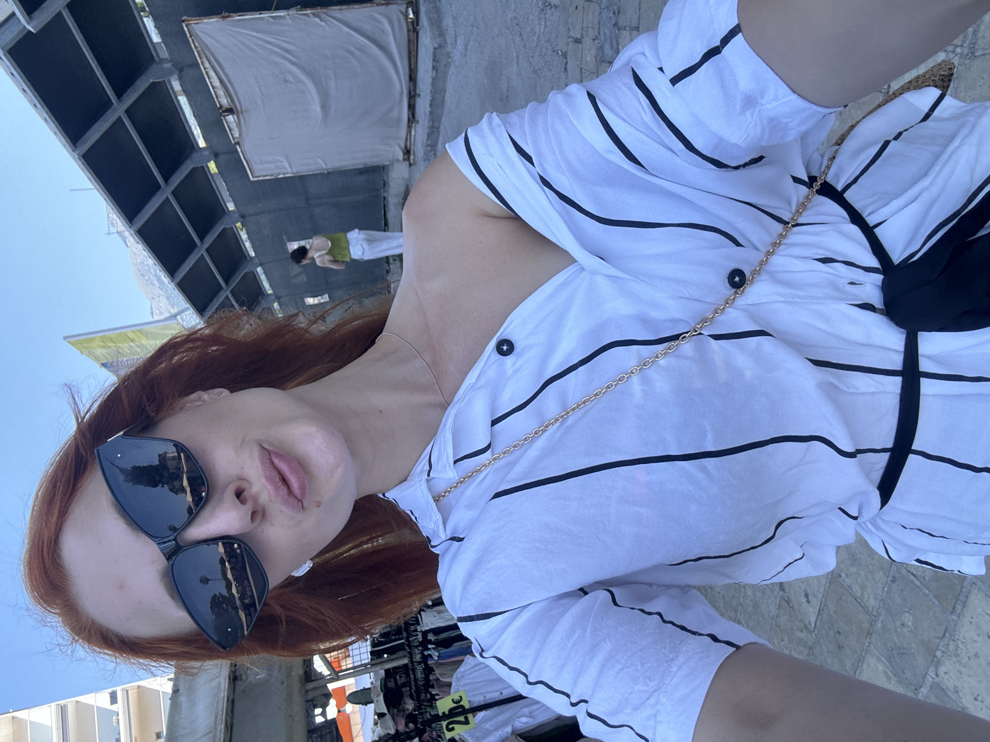 A woman with red hair is taking a selfie outdoors. She is wearing sunglasses and a white and black striped shirt, with a gold chain bag strap visible across her chest. The background appears to be a street or market, with some stalls and buildings visible. There is a price tag with "2€" written on it. The scene is brightly lit, suggesting it is daytime. The overall impression is casual and relaxed.