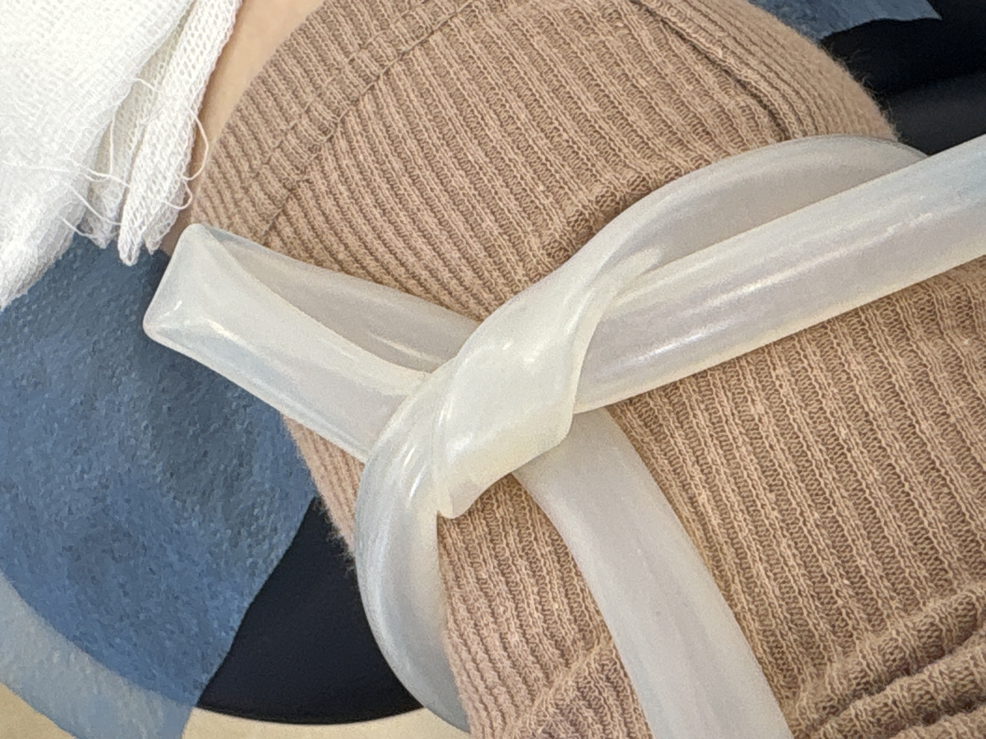 This close-up image captures a detail of what appears to be a medical or clinical setting. The central focus is a semi-transparent, milky-white, flexible tube or strap, possibly a tourniquet, wrapped around a limb, which is largely covered by fabrics.
The limb is obscured by a light brown, ribbed, knitted fabric, likely a sleeve of clothing or a blanket, which occupies a large portion of the frame. Overlapping this and situated towards the upper left is a piece of white, loosely woven gauze or bandage, showing visible threads. To the left, a blue fabric, possibly a medical drape or part of a bedsheet, is visible, contrasting with the beige and white materials. A small section of dark blue or black fabric is also seen towards the lower left.
The translucent tube is twisted and looped, creating a knot-like appearance where it crosses itself over the beige fabric. The lighting suggests an indoor environment, possibly under artificial light. There is no visible activity other than the static arrangement of these items, suggesting preparation for or the aftermath of a medical procedure such as a blood draw or IV insertion. No people are visible, nor is any text present in the image. The provided context indicates the location is Saint Petersburg, Russia.