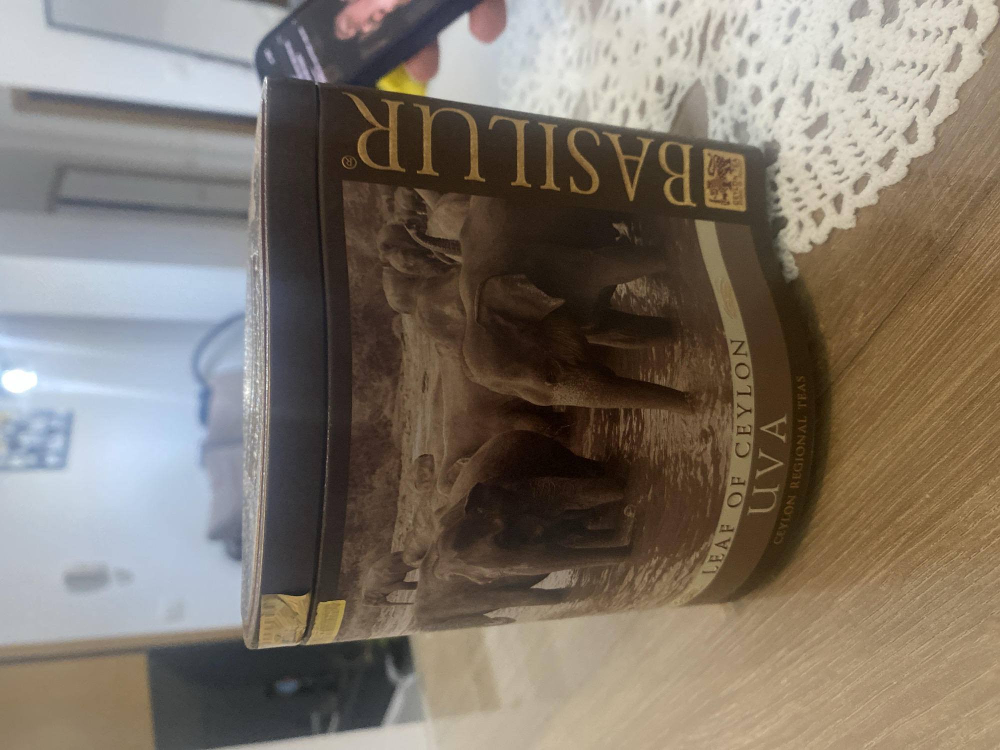 Here is a description of the image:
The image shows a cylindrical tin of Basilur brand Ceylon tea, specifically the UVA variety. The tin's design features a sepia-toned image of elephants wading in water. The label also displays the words "LEAF OF CEYLON UVA" and "CEYLON REGIONAL TEAS." The location of the scene is unknown.