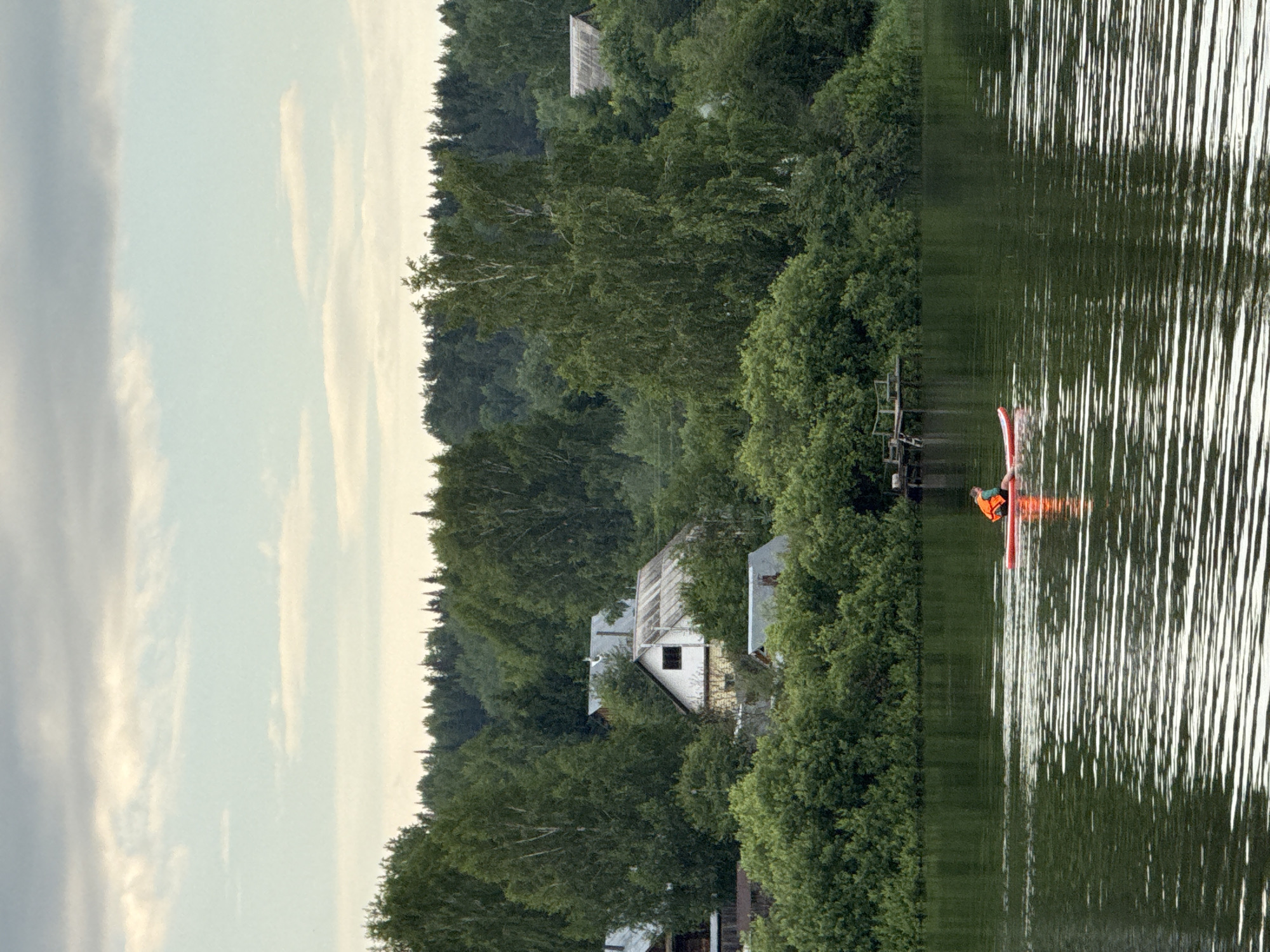 In Luzhkovo, Russia, a person wearing an orange life vest paddles a red stand-up paddleboard across a calm, dark green body of water. The background features a dense line of lush green trees, behind which are several houses with gray, peaked roofs, nestled among the foliage. The sky above is overcast with soft, muted clouds.