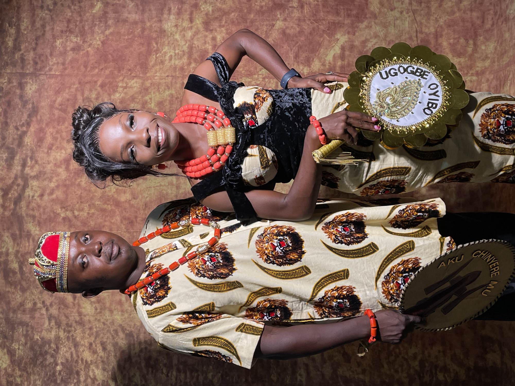 Two individuals, a man and a woman, stand side-by-side in traditional Nigerian attire, posing for a photograph against a mottled brown and gold backdrop. The scene suggests a cultural celebration or formal event, likely in Jos, Nigeria.
The woman, on the viewer's right, smiles broadly at the camera. She wears a black velvet top and a flowing skirt or wrapper made of a light cream-colored fabric patterned with lion-like heads and gold curved shapes. Her hair is styled in an updo, and she is adorned with multiple strands of large, vibrant coral red beads around her neck, featuring an ornate gold centerpiece, and a matching bracelet. She holds a circular golden fan decorated with a white center and gold embroidery. Visible text on her fan reads "UGOGBE OYIBO".
The man, on the viewer's left, also smiles gently at the camera. He wears a short-sleeved shirt made from the same patterned cream fabric as the woman's bottom garment. He has a single strand of coral beads around his neck and a matching bracelet on his left wrist. On his head, he wears an elaborate red and gold crown-like cap, intricately decorated with beads and sequins. He holds a round, brown fan or shield-like object with raised designs and text that reads "AKU CHINYERE MU".
Both individuals appear joyful and proud, reflecting the celebratory nature of the event. Their elaborate clothing and accessories, particularly the coral beads, are characteristic of Igbo cultural attire.