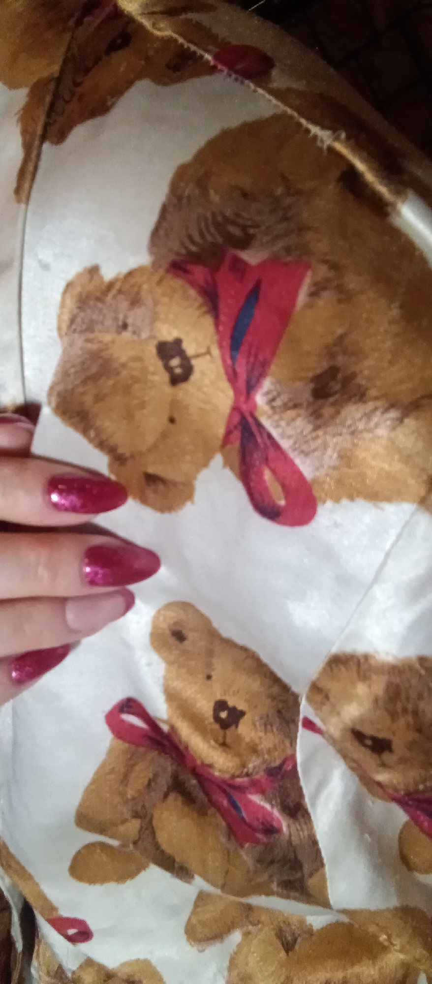 The image presents a close-up view of a person's hand with long, pointed fingernails, distinctively painted with a shimmering, dark pink or magenta glitter polish. The hand is positioned against a light-colored, possibly white, fabric that serves as the background. This fabric is adorned with a repeated pattern of classic brown teddy bears. Each teddy bear in the pattern is depicted with dark eyes and nose, and wears a red bow tied around its neck; some bows also show subtle blue stripes. The fabric appears soft and somewhat creased, suggesting it could be part of bedding, a blanket, or an item of clothing like pajamas. The scene is an intimate, indoor setting, with no specific time of day or weather discernible. No particular activity is taking place beyond the display of the hand and the patterned fabric. There is no visible text in the image.