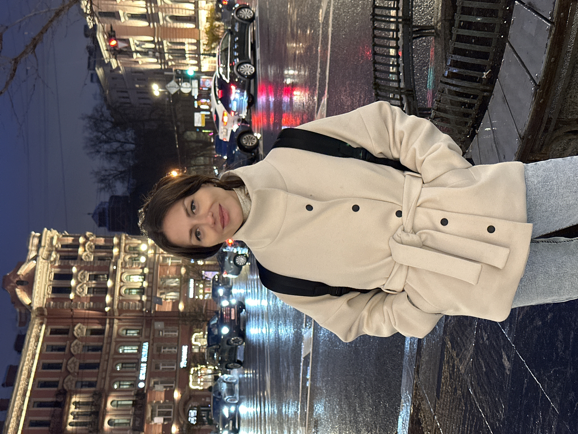 A young woman stands outside on a dark, rainy night in Saint Petersburg, Russia. She is wearing a light-colored coat with a belt and a black backpack. The street behind her is wet, reflecting the lights of passing cars. In the background, a large, ornate building is illuminated with lights. The reflections on the wet pavement add to the city's nighttime atmosphere.
