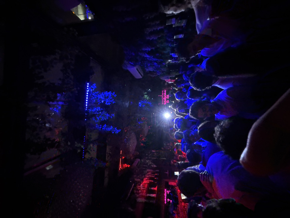 The image captures a densely crowded indoor space resembling a nightclub or bar, characterized by low-level, multi-colored lighting during evening hours. The environment is predominantly dark, illuminated by ambient blue and red light sources. Overhead and along walls, artificial green foliage, some adorned with blue string lights, forms part of the decor. A row of blue LED strip lights is visible on the upper ceiling. Red lighting fixtures are present on the left side of the frame. A bright white spotlight emanates from the background, center. An air conditioning unit is mounted on the upper right wall. In the background, a pink neon sign is partially legible, displaying "Welcome to Th...".
Numerous individuals, appearing to be young adults, are tightly packed throughout the foreground and midground. They are standing close together, engaging in social interactions or dancing. Their attire largely consists of light-colored shirts that reflect the colored ambient lighting. On the far left, one person is seen with an arm around another. Most faces are obscured by shadows, backlighting, or other individuals.
The specific location within Washington, United States, cannot be identified from the visual evidence in the image.
