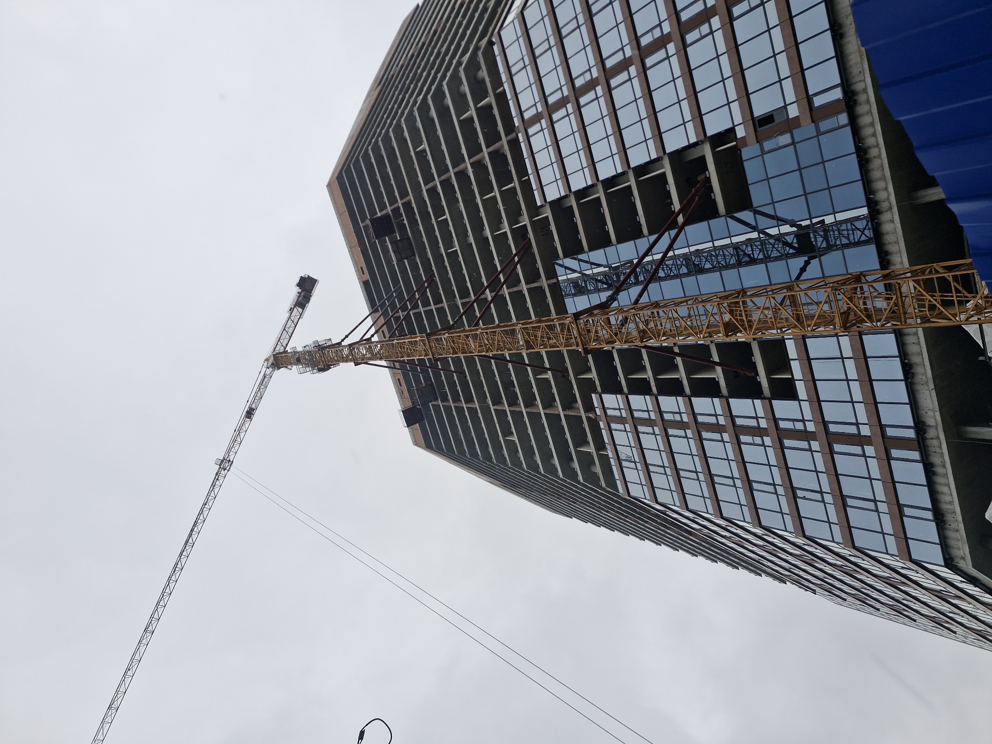 A low-angle shot depicts a building under construction with a large crane in the foreground, set against an overcast sky. The unfinished building has sections with windows installed and others where the concrete frame is exposed. A yellow construction crane stands adjacent to the building, extending its boom outwards. The sky is predominantly gray and featureless. This is likely a construction site in Yekaterinburg, Russia.