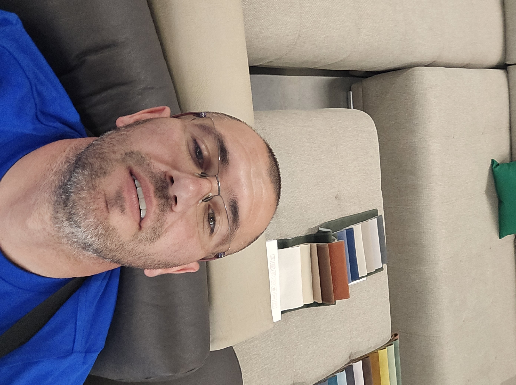 The image is oriented 90 degrees counter-clockwise, showing a man reclining on a sofa, looking directly at the camera.
**Subjects:** The primary subject is a male, likely middle-aged, with light skin, short dark hair, and visible gray stubble on his face. He is wearing glasses with thin metal frames and a bright blue t-shirt. His expression is neutral.
**Objects:** The man is situated on a sofa, which appears to be a sectional piece. One section where he is leaning is dark gray or dark brown, while the larger background sections are upholstered in a light beige or tan fabric. On the light-colored sofa section to his right, there are several fabric swatch books or sample cards arranged. These samples display various colors and textures; one visible set includes warm tones like beige, brown, and burnt orange, while another partially visible set appears to have blues, grays, and whites. A small, bright green cushion or pillow is visible in the background, resting on the sofa.
**Scene and Setting:** The scene is indoors, strongly suggesting a furniture store or showroom, given the presence of multiple fabric samples and the modular appearance of the sofa.
**Activity/Event:** The man appears to be either resting or posing for a picture on the sofa, possibly in the process of selecting furniture or fabric options.
**Notable Details:** The image's orientation places the man's head to the left side of the frame. No specific time of day or weather can be determined. There are some indistinct small icons visible on one of the fabric sample cards, but no discernible text or brand names.