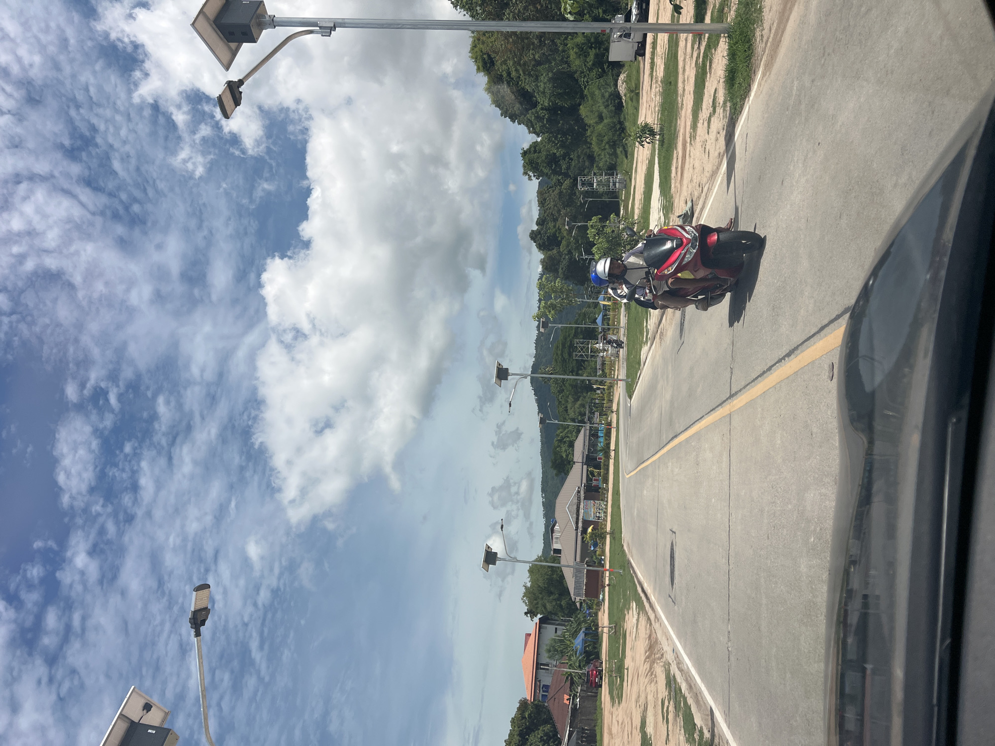 In a sunlit outdoor scene in Ban Bang Rak, Thailand, a two-lane paved road curves towards the right. Two people are riding a red scooter, with the driver wearing a blue helmet and the passenger wearing a white helmet. The road is marked with a yellow line down the center and a white line on the left edge. Solar-powered streetlights are visible on poles along the roadside. On the left side of the road, there are some buildings with red and orange roofs, as well as a blue tent. On the right side, there are trees, grass, and a white truck. The sky is blue with scattered white clouds. The photo appears to be taken from inside a vehicle, with the dashboard visible in the foreground.