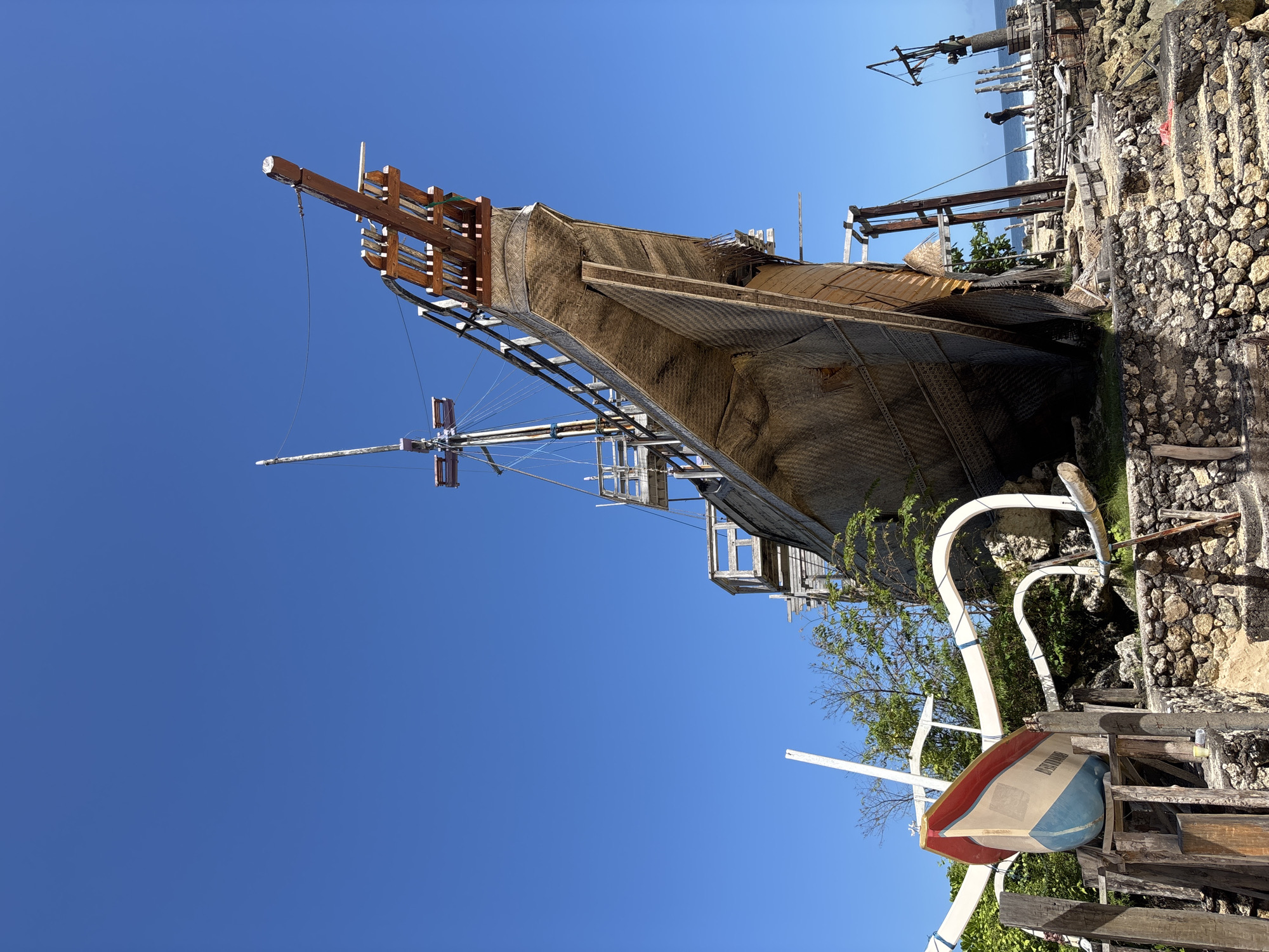 The image shows a large, decaying wooden structure resembling a ship's hull in Kuta Selatan, Indonesia. The exterior of the hull is covered in woven material. Various wooden scaffolding and supports surround the structure, with ropes visible. Part of a smaller, overturned boat painted white, blue, and red is in the foreground. Rocky terrain surrounds the structure. The sky above is clear and blue. In the background, a glimpse of the ocean can be seen.