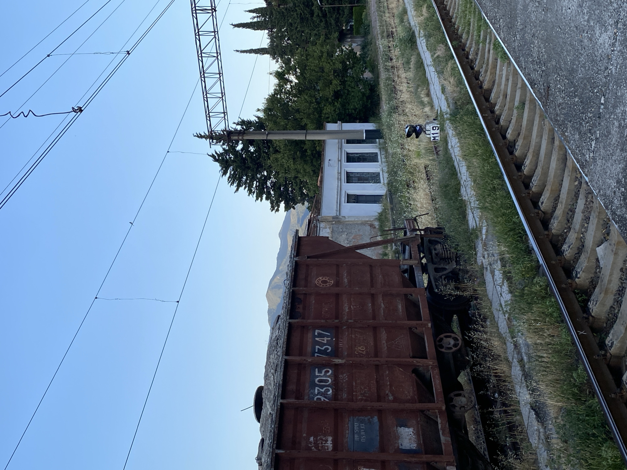 This image captures a train track area with a freight car in the foreground. The train car is rusty and appears to be an older model, with the numbers "2305 7347" visible on its side. It is parked on a track lined with concrete sleepers and gravel. Beyond the tracks, there is a low, white building with multiple windows and what appears to be a signal light labeled "M19" positioned near the tracks. Several tall, green trees stand behind the building, and in the distance, mountains are faintly visible. Overhead, power lines and a metal support structure for them dominate the upper left portion of the frame, against a clear blue sky. The lighting suggests it is daytime, likely late afternoon or early morning, given the soft light. The scene appears to be a quiet, perhaps disused, section of a railway line in Mtskheta, Georgia, with no people visible. The overall impression is one of stillness and a sense of a bygone era due to the aged appearance of the train car and the infrastructure.