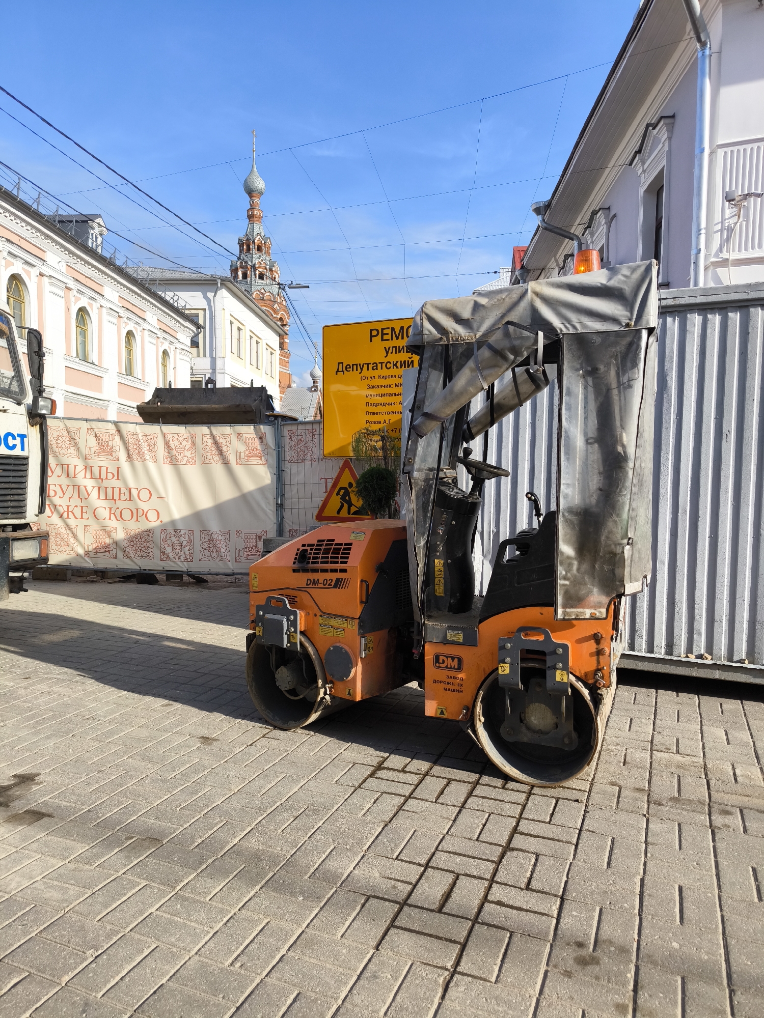 An orange road roller is parked on a paved sidewalk in front of a white corrugated metal wall and a temporary construction banner. The banner displays red decorative patterns and text that reads "УЛИЦЫ БУДУЩЕГО - УЖЕ СКОРО." ("Streets of the future - coming soon."). To the left, part of a white truck is visible. Behind the roller, a yellow sign with Russian text is mounted on a pole. The sign reads "РЕМОНТ улиц Депутатский..." ("Repair of streets..."). In the background, a multi-tiered red and gray church tower with a gold cross on top pierces the clear blue sky. The buildings lining the street have cream-colored facades with arched windows. Overhead, a network of dark electrical wires crisscrosses the sky. The sun casts long shadows on the brick-paved ground.