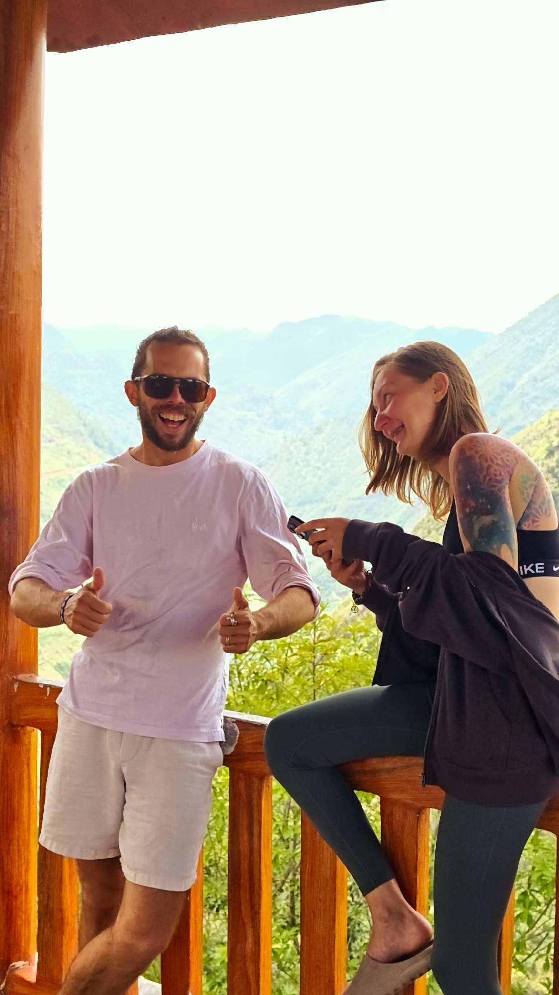 A man and a woman are standing on a wooden veranda in what appears to be the city of Lũng Cú Commune, Vietnam, given the mountainous landscape in the background.
The man on the left has dark hair and a beard, and is wearing sunglasses, a light purple long-sleeved shirt, and white shorts. He is smiling and giving two thumbs-up.
The woman on the right is smiling and looking at something in her hands, possibly a phone or other small device. She has short brown hair and a tattoo on her upper left arm. She is wearing a black sports bra with "NIKE" printed on it, dark green leggings, and slip-on shoes. She is also wearing a dark jacket. The veranda has wooden railings. The background reveals a mountainous, somewhat hazy landscape, with green vegetation visible. The photo is well-lit, with natural light.