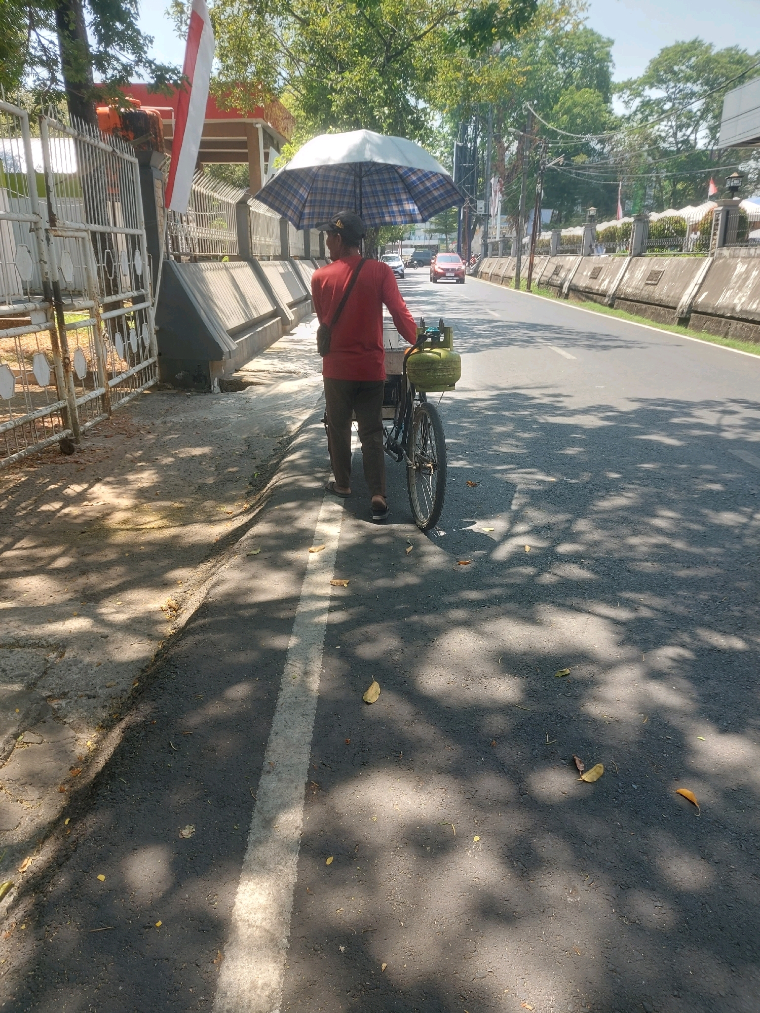 The image depicts a person walking with a bicycle on a paved road, holding an open umbrella overhead. The person is wearing a red shirt and dark pants, and their back is to the camera. The bicycle has a green gas cylinder attached to it. The scene is set in a city, with buildings and trees visible in the background. The road has a white line marking the lane. Sunlight is casting shadows on the road and sidewalk. The presence of a gas cylinder on a bicycle suggests a delivery or vending activity. Based on the provided context, this scene is located in the city of Makassar, Indonesia, likely on a street with sidewalks and traffic.