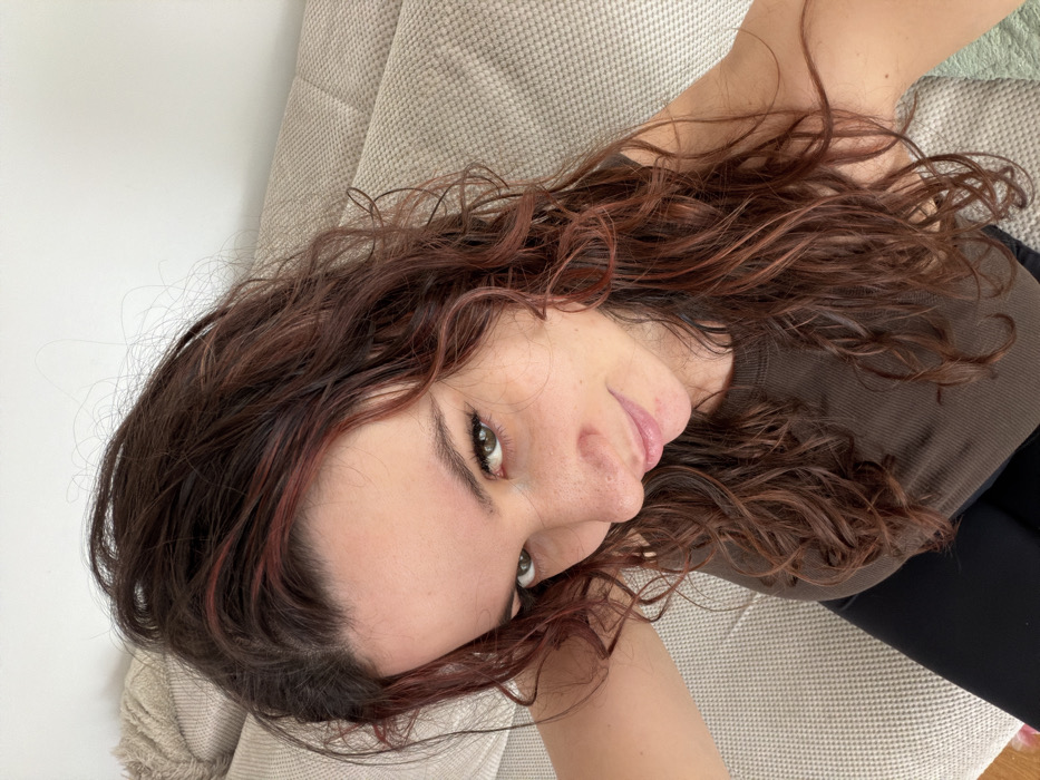 The image displays a close-up, high-angle self-portrait of a single female individual indoors. The subject has long, wavy, dark brown hair with reddish-auburn highlights, which appears slightly damp. Her pale complexion is visible, with freckles across her nose and cheeks. She has green-brown eyes, accentuated with eyeliner, looking directly into the camera with a subtle smile. She is wearing a ribbed, dark brown tank top or crop top, and black clothing is visible below her waist. The background features a light-colored, textured upholstered sofa or couch to the subject's left and a plain white wall in the upper right. The specific location in the city of Budva, Montenegro, cannot be identified from the visual content.
