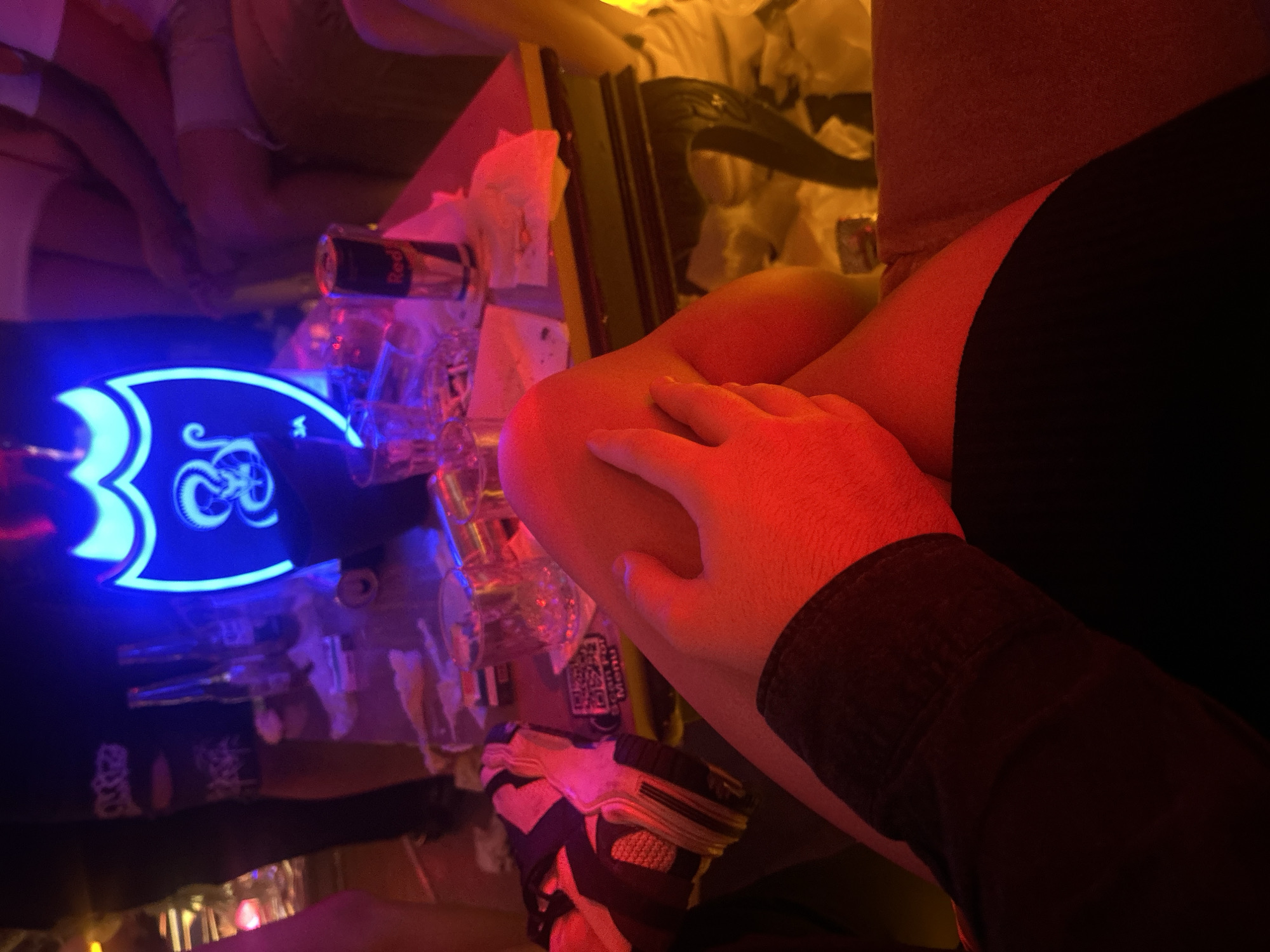 The image appears to be taken inside a bar or nightclub in Pleven, Bulgaria. There are people seated around a table with multiple empty shot glasses and a Red Bull can. A blue neon sign with a stylized logo is visible. A person's hand rests on the arm of another individual wearing a dark shirt. A partial view of a sneaker is also present. The scene is illuminated by colored, likely red and blue, lighting.