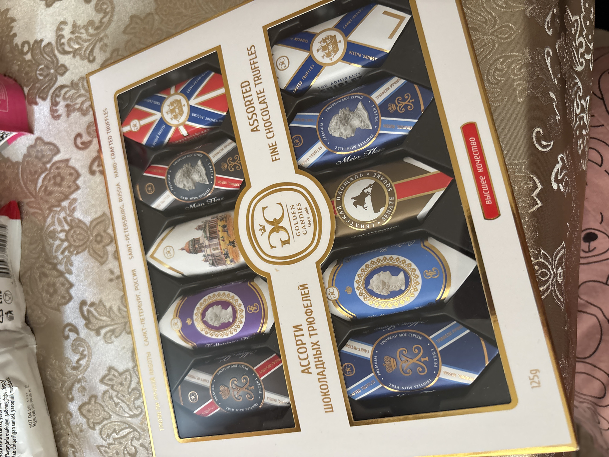 This media file is a close-up, top-down shot of an opened box of assorted fine chocolate truffles. The box is white with gold trim and is filled with twelve individually wrapped truffles arranged in two rows of six. The truffles have decorative wrappers featuring various designs, including the Union Jack, a regal portrait, and an architectural building. Some wrappers also have Russian text. The text "ASSORTED FINE CHOCOLATE TRUFFLES" is printed vertically on the left side of the box, and "SAINT-PETERSBURG, RUSSIA HAND-CRAFTED TRUFFLES" is printed horizontally along the side. On the right side, there is a red label with white Russian text that reads "Высшее качество" (Highest Quality), and "125g" is printed on the bottom right corner of the box. The box is placed on a textured fabric surface with a decorative pattern, possibly a tablecloth or blanket. The overall impression is one of a well-presented gourmet product.