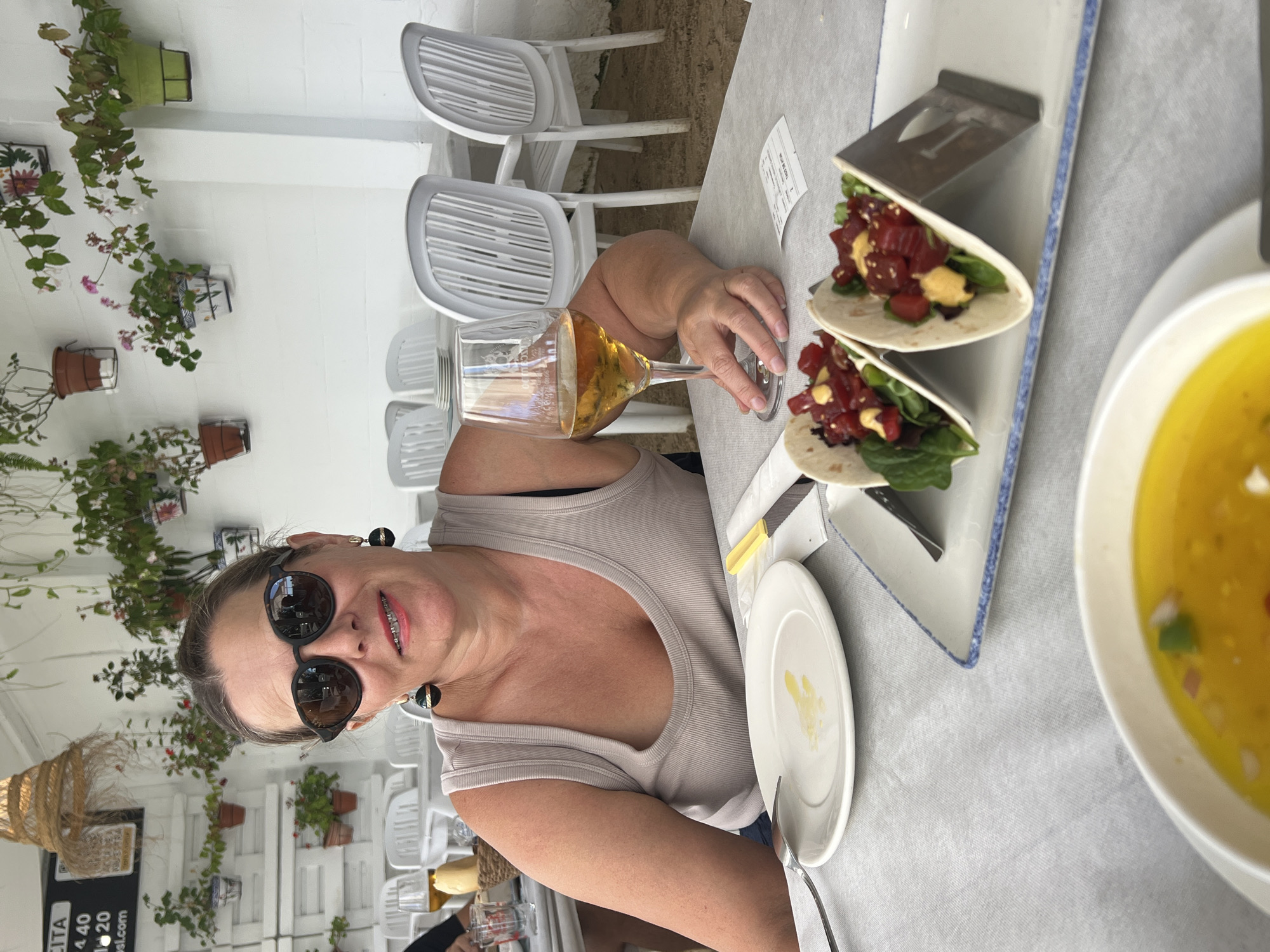 A woman with sunglasses and a tank top sits at a table in an outdoor restaurant, possibly in Valdeconejos, Spain. She is holding a glass of amber-colored liquid. In front of her are two tacos, a white plate with some yellow sauce, and a bowl of yellow liquid with diced red and green ingredients. White plastic chairs and a white wall decorated with potted plants are visible in the background. The scene appears to be a meal at a casual dining establishment.