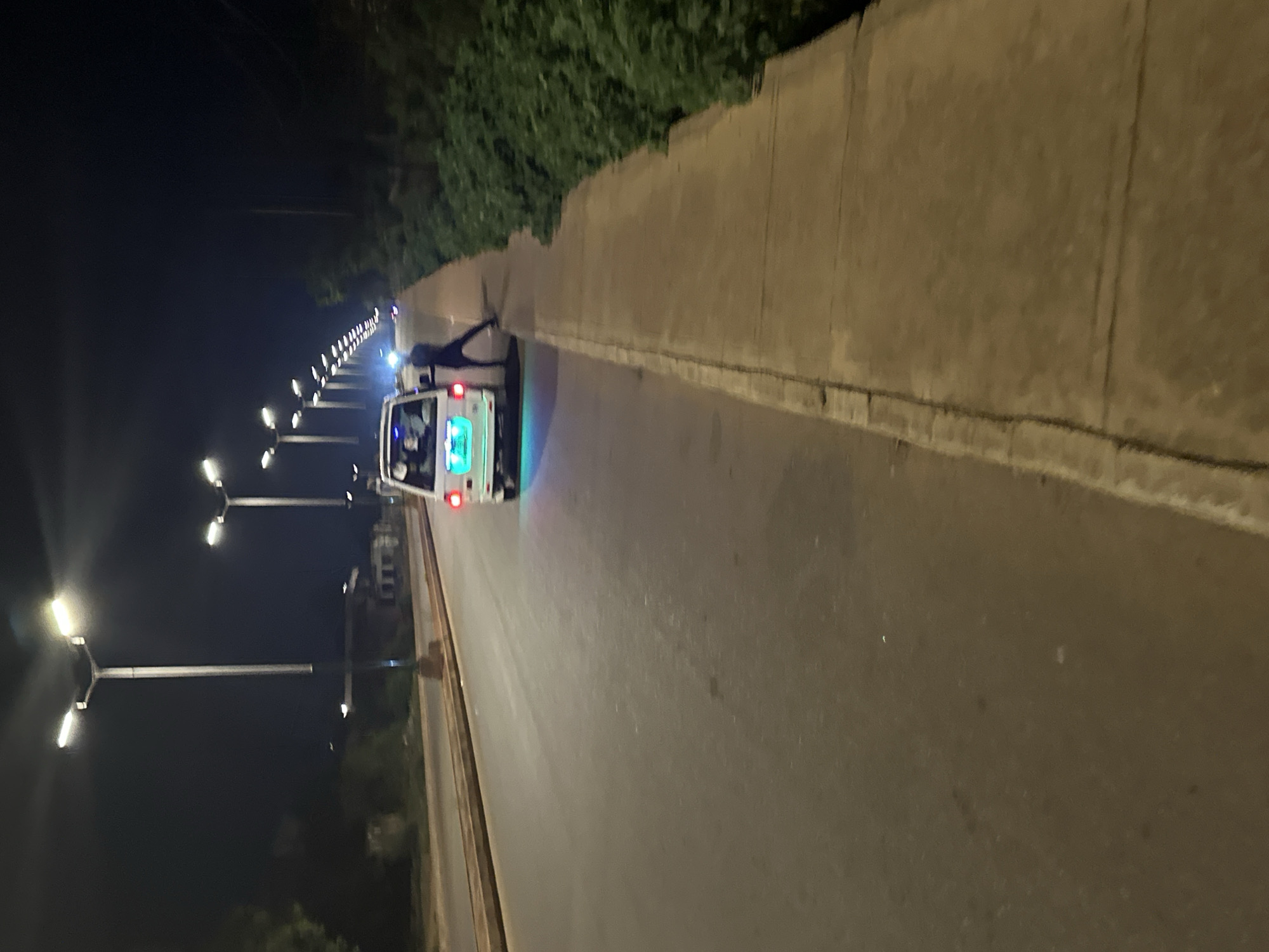 The image shows a lit road at night, possibly in Bukuru, Nigeria, though there is no way to verify this. A white van with illuminated lights is moving along the road. A person is standing next to the vehicle. Streetlights line the left side of the road. A concrete barrier and vegetation are present on the right side. The overall environment is dark, with the scene illuminated by streetlights and vehicle lights.