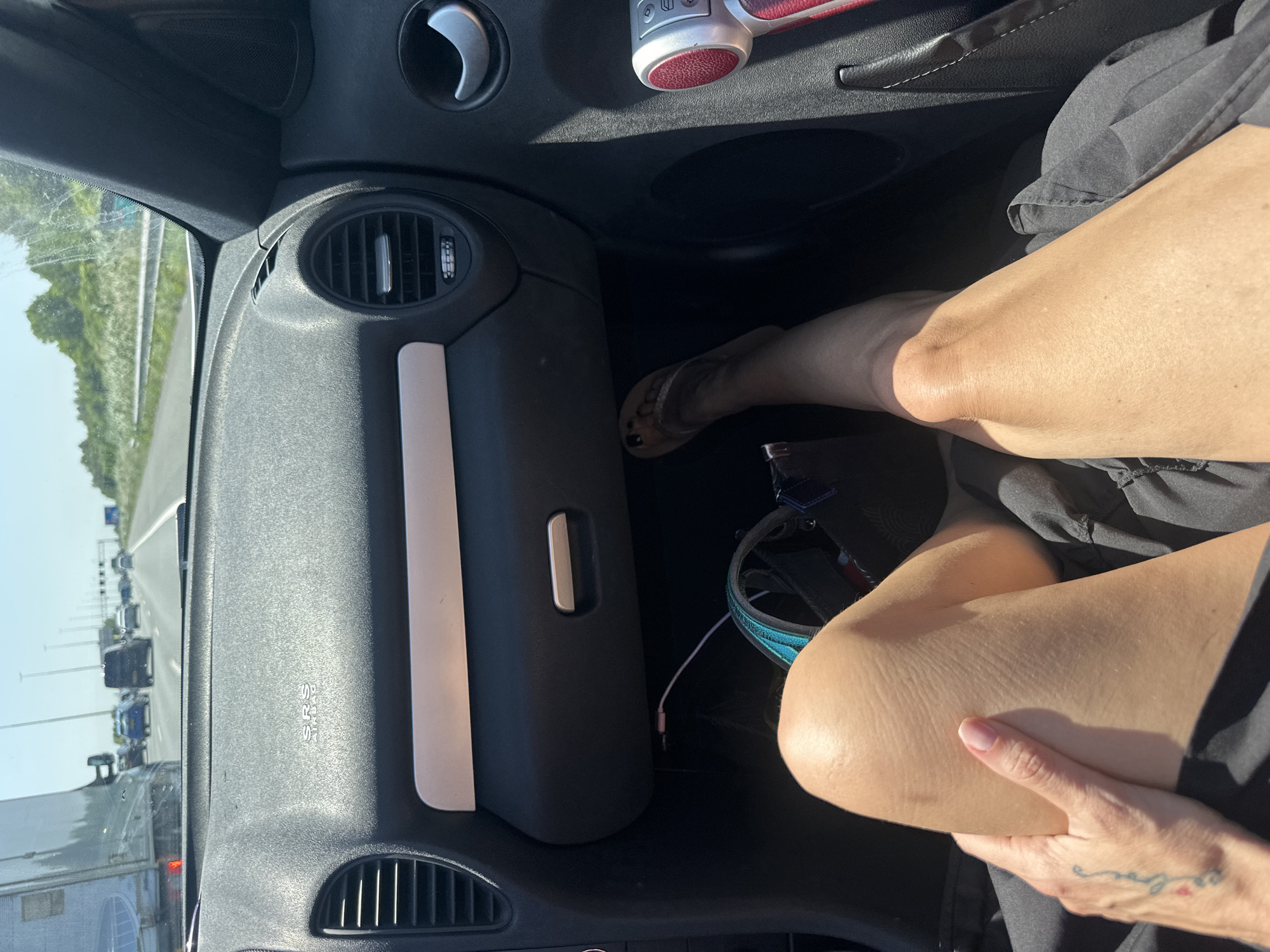 The image depicts an interior view from the passenger side of a vehicle, presumably a car, situated on a highway in Leidschendam, Netherlands, as evidenced by the traffic congestion visible through the windshield. The driver is absent from the frame, and a passenger's legs are resting towards the dashboard area, with the knees bent and a hand visible. The dashboard features ventilation systems and an SRS (Supplemental Restraint System) indicator. A pair of flip-flops rests near the passenger's foot. A black bag and an electronic cable are present in the space near the passenger's legs.