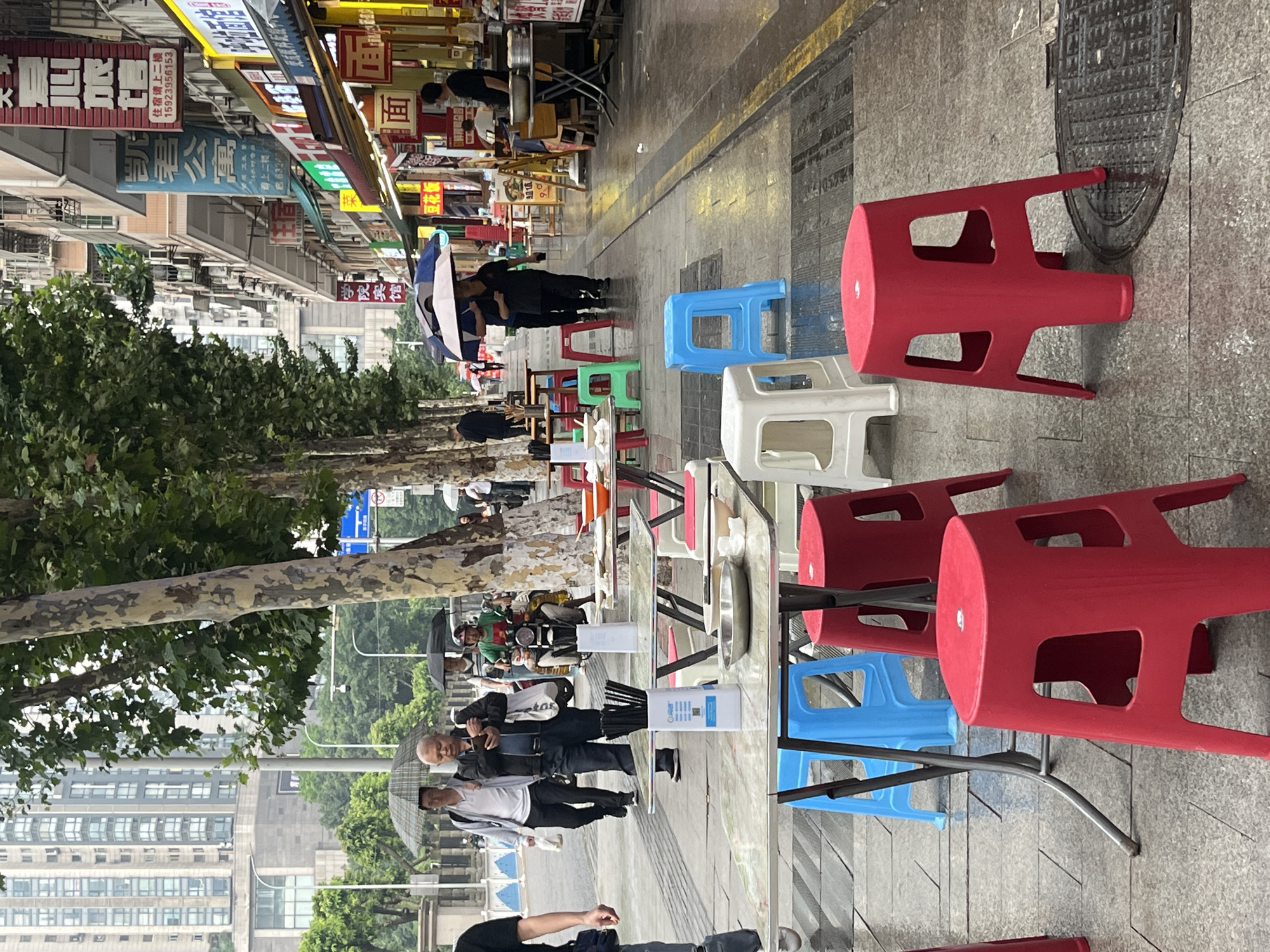 The image depicts a street scene in Yuzhong District, China, likely a commercial area with shops and eateries lining the sidewalk. The weather appears to be rainy or recently rained, as evidenced by the wet pavement and several people holding umbrellas.
In the foreground, several plastic stools of various colors (red, blue, white) are placed around tables set up for outdoor dining. These tables are covered with patterned tablecloths, and some have bowls and chopsticks on them, suggesting an active food service.
Further back, pedestrians are walking along the sidewalk, some carrying umbrellas. One man is holding a mobile phone and looking down at it. A motorcyclist is visible in the middle ground, also with an umbrella. Shops with prominent signage, some featuring Chinese characters, are visible on the right side of the street. Trees with textured bark line the left side of the street, providing some shade. In the background, modern high-rise buildings indicate the urban setting. The overall atmosphere is that of a bustling city street, adapted for outdoor dining despite the weather.