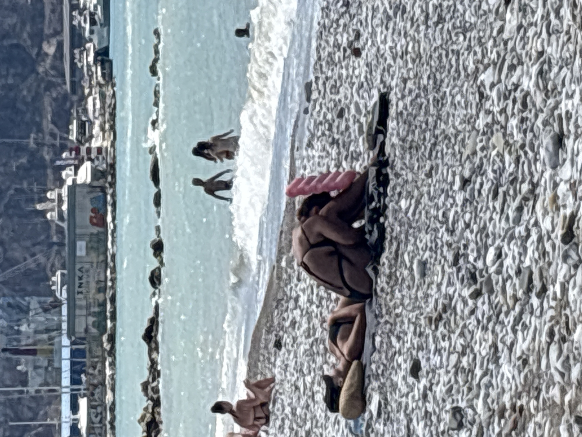 The image depicts a crowded beach scene in the city of Bar, Montenegro. In the foreground, two women are sunbathing on the pebbled shore. One woman is lying on her back, with a pillow under her head, and is wearing a black bikini. The other woman is sitting and partially obscured by a pink object, also wearing a bikini.
In the middle ground, several people are wading and playing in the shallow water. Three individuals, appearing to be adults, are visible among the breaking waves. Further out in the sea, a person is visible in the water.
The background reveals a coastal landscape with a rocky breakwater separating the beach from the sea. Beyond the water, a port area is visible, with various boats and yachts docked. Buildings, including one with the word "INKA" graffiti on it, are also present in the port. Mountains form the backdrop to the entire scene. The water is a vibrant turquoise color, and the sky is overcast.