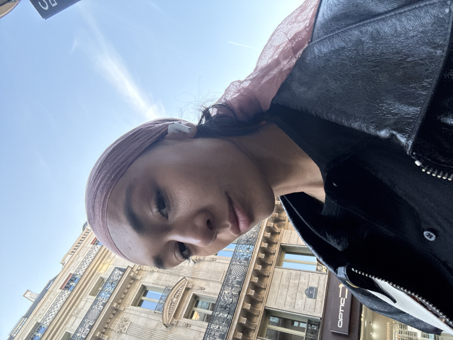 A close-up photograph features an individual, appearing to be a female adult, positioned in the foreground and facing the camera directly. She has medium skin tone and wears a light pink, semi-sheer headscarf, a dark shirt, and a black leather-like jacket with a prominent silver zipper. A white wireless earbud is visible in her right ear. Her expression is neutral, with slightly parted lips.
The background reveals a multi-story building of classic Haussmannian architecture, constructed from light-colored stone, featuring ornate wrought-iron balconies and tall windows. A street sign visible on the building identifies its location as "RUE SAINTE-ANASTASE," placing the scene in Paris, France. Commercial establishments are present at street level, including a store labeled "OPTIC" and a "BRASSERIE." The sky overhead is clear blue with minimal, faint contrails. The scene is illuminated by natural daylight.