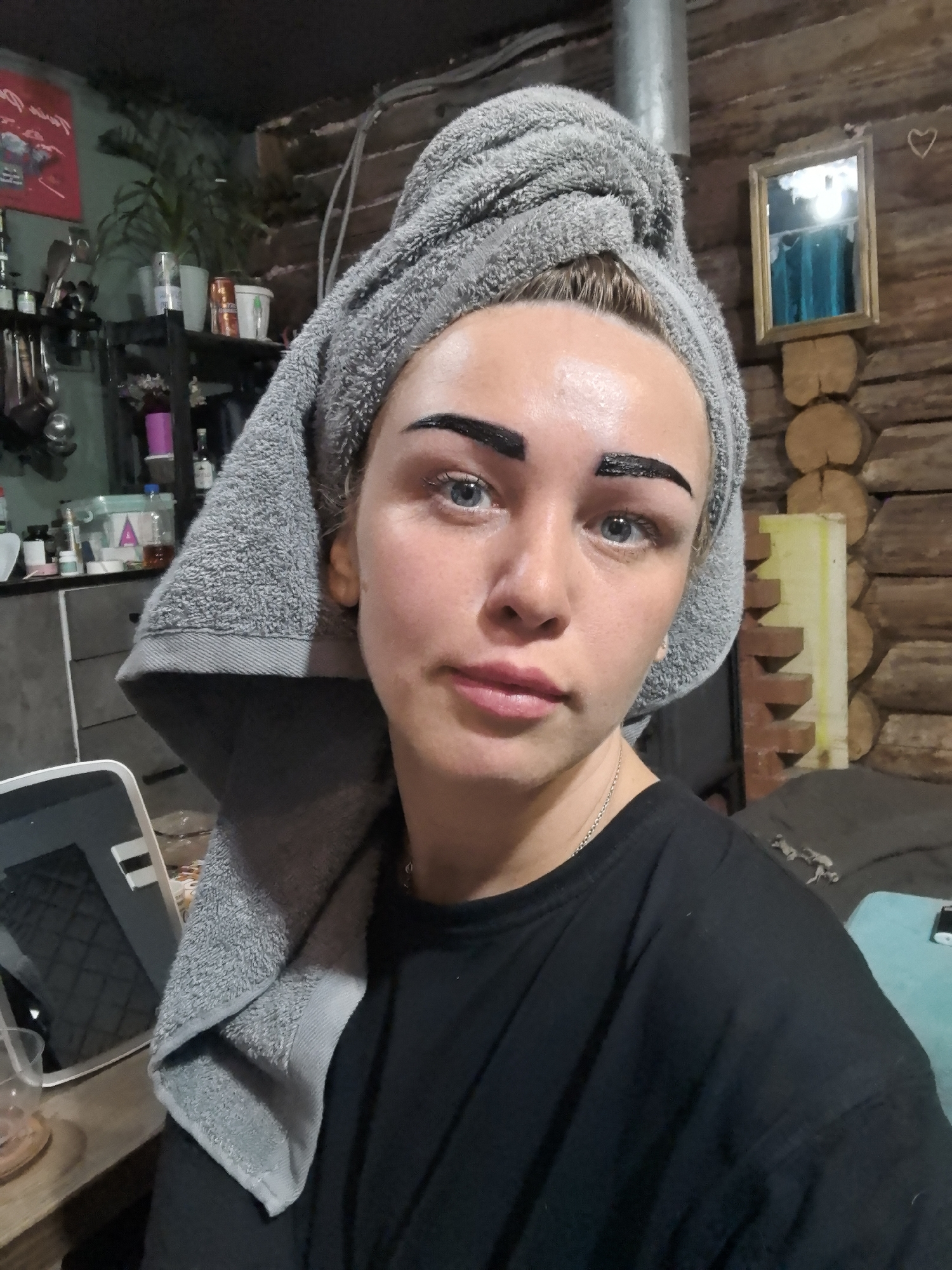 A woman is pictured indoors, wearing a grey towel wrapped around her head. Her eyebrows are coated with a dark substance, likely dye or henna. She is fair-skinned, with blue eyes and pink lips, wearing a black t-shirt and a silver necklace.
The background includes a cluttered shelf with various items, a log cabin wall, and a mirror. The location cannot be confirmed to be in the city of Noviy, Russia.