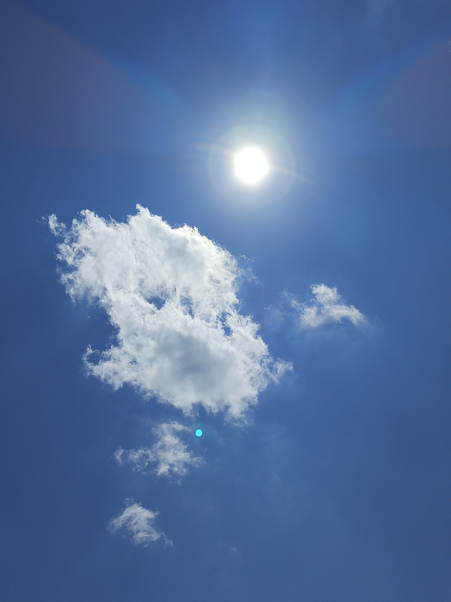 A bright, white sun with visible rays dominates the upper portion of a clear blue sky. A large, fluffy white cloud is positioned below and slightly to the left of the sun. To the right of this main cloud, there are several smaller, wispy clouds. A small, bright turquoise dot is visible in the sky to the lower right of the large cloud, and another smaller white cloud is present at the bottom left of the image. The image appears to be taken from a ground-level perspective looking upwards.