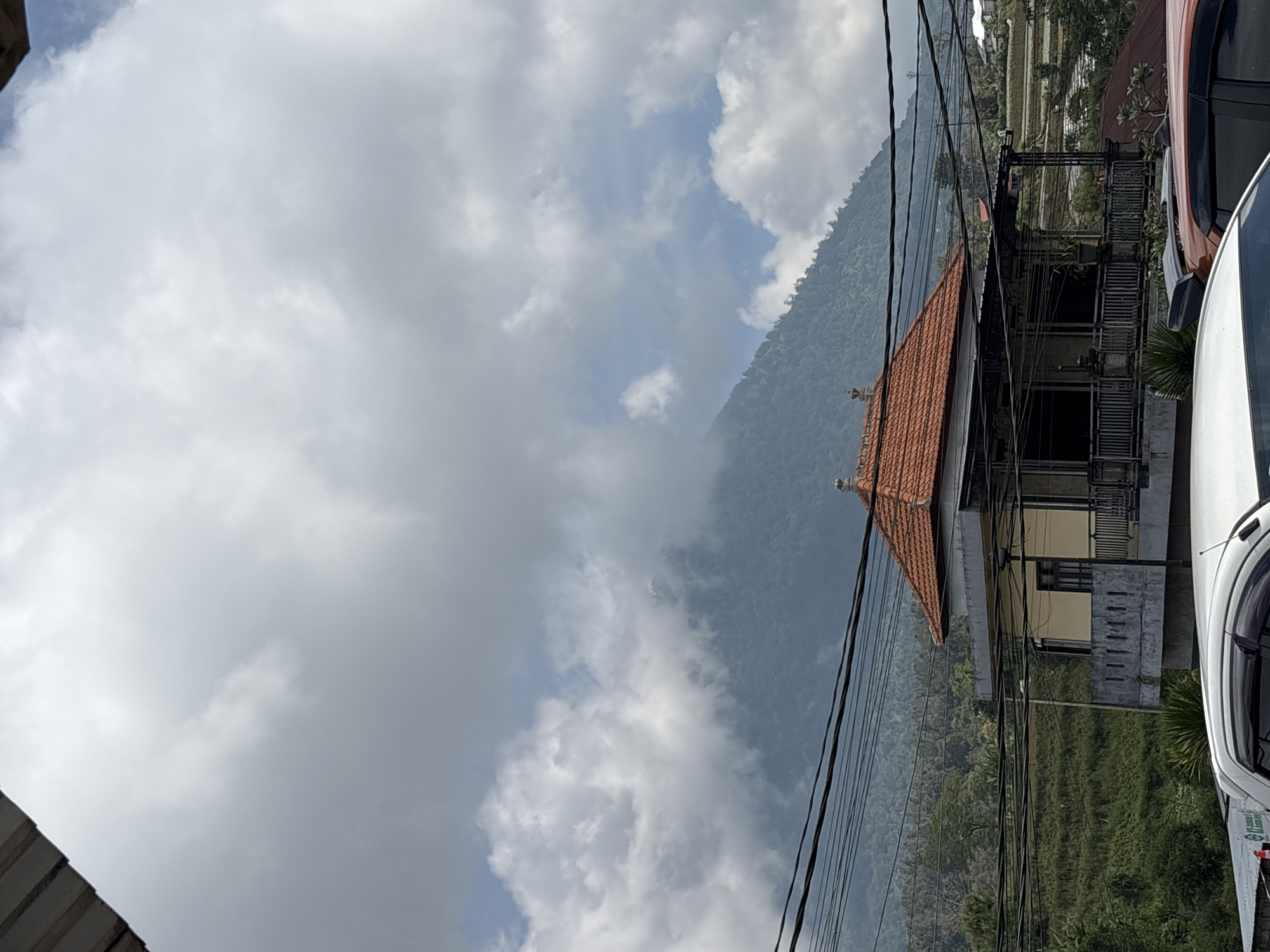 The image depicts a partially obscured scene in Baturiti, Indonesia, notable for its overcast sky and mountainous terrain. A building with a red-tiled roof is visible, partially hidden by utility lines. Adjacent to the building is a white vehicle. The surrounding landscape is green with vegetation, suggesting a lush environment. Terraced rice fields are also discernible in the background. The perspective is angled, and details are limited due to the obstruction of the power lines and partial view.