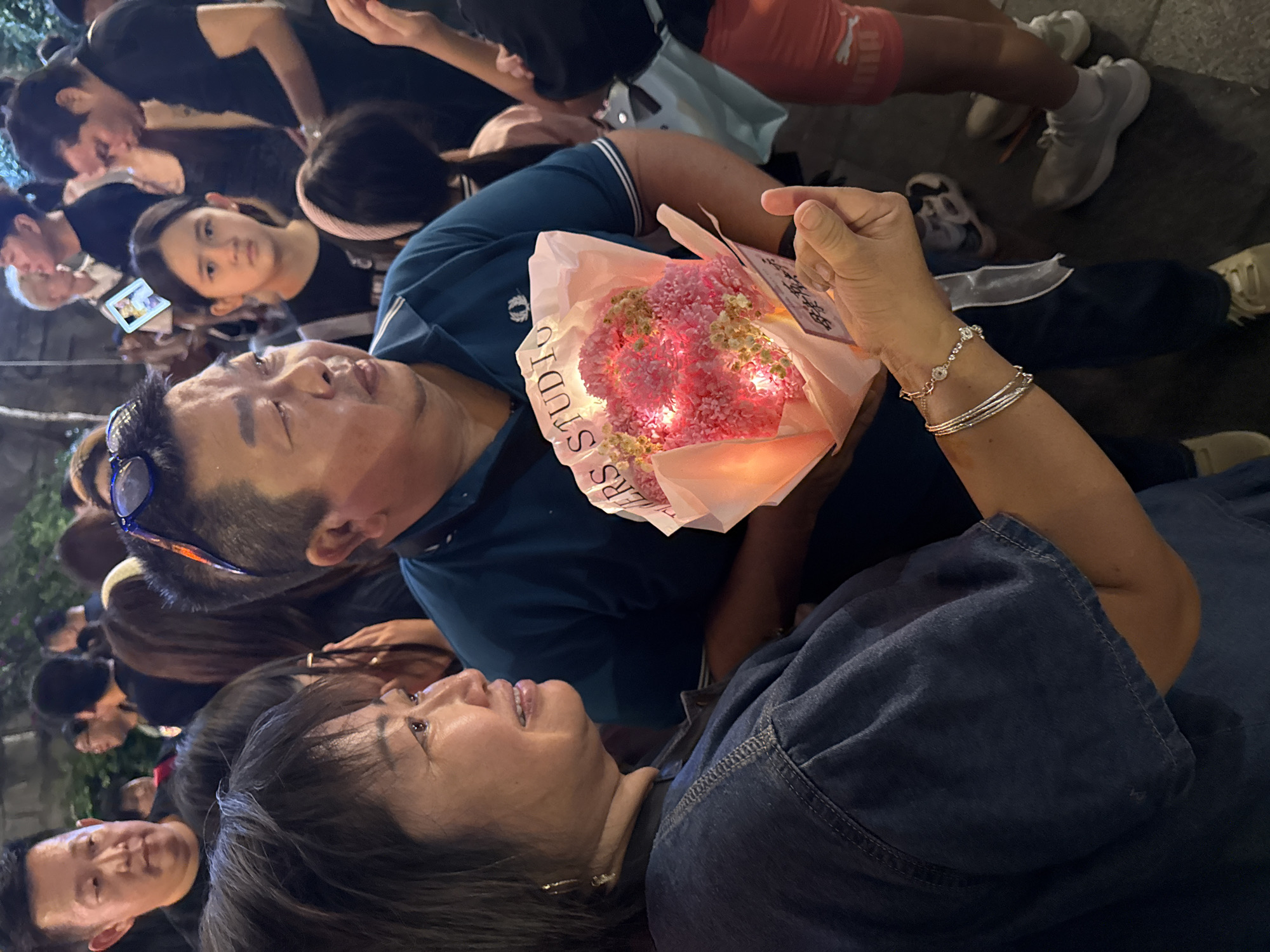 A group of people are gathered together, with a woman holding up a bouquet of flowers that appear to have pink lights intertwined within them. The woman is wearing a denim shirt or jacket, and several silver bracelets are visible on her wrist. A man in a blue polo shirt is looking at the flowers as well. A young girl standing behind them is looking directly at the camera. Others in the background are also observing the scene, including another man in the lower left corner. A second woman can be seen behind the bouquet. The scene takes place outdoors. The image appears to have been taken in Yuzhong District, China.