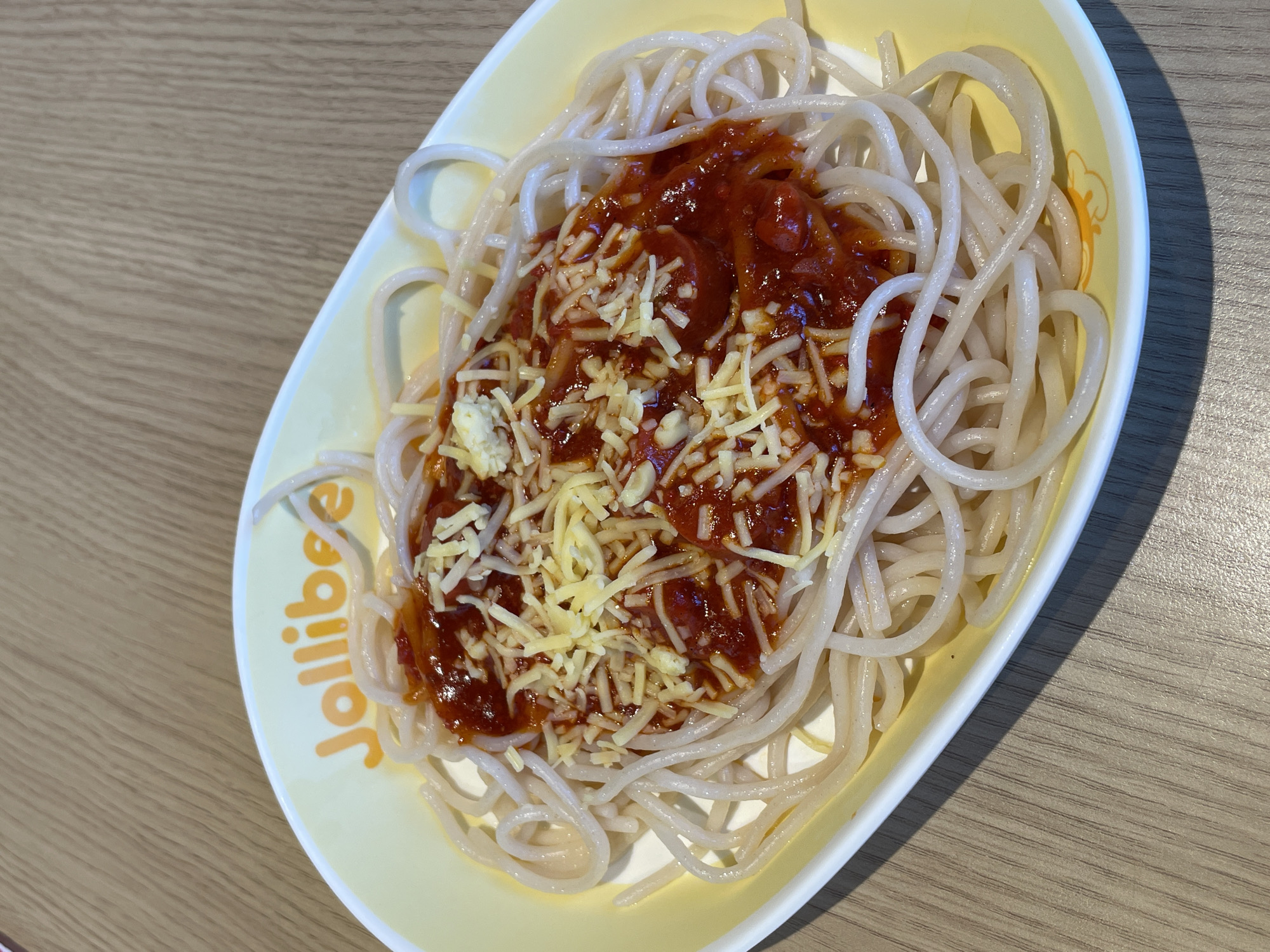 A close-up, overhead shot shows a Jollibee spaghetti dish on a light wood table. The plate, a pale yellow oval with the Jollibee logo in orange, contains a generous portion of spaghetti noodles covered in a red meat sauce and topped with shredded cheese. The noodles are long and thin, with a slight sheen. The sauce is thick and appears to have chunks of meat and possibly hotdog. The cheese is finely grated and sprinkled unevenly over the sauce. The background shows the texture of the wood grain on the table, with a shadow cast by the plate on the right side of the image. The lighting is bright and even, suggesting it was taken indoors in a well-lit environment. There are no people visible in the image. This scene is consistent with a meal at a Jollibee restaurant in Davao City, Philippines.