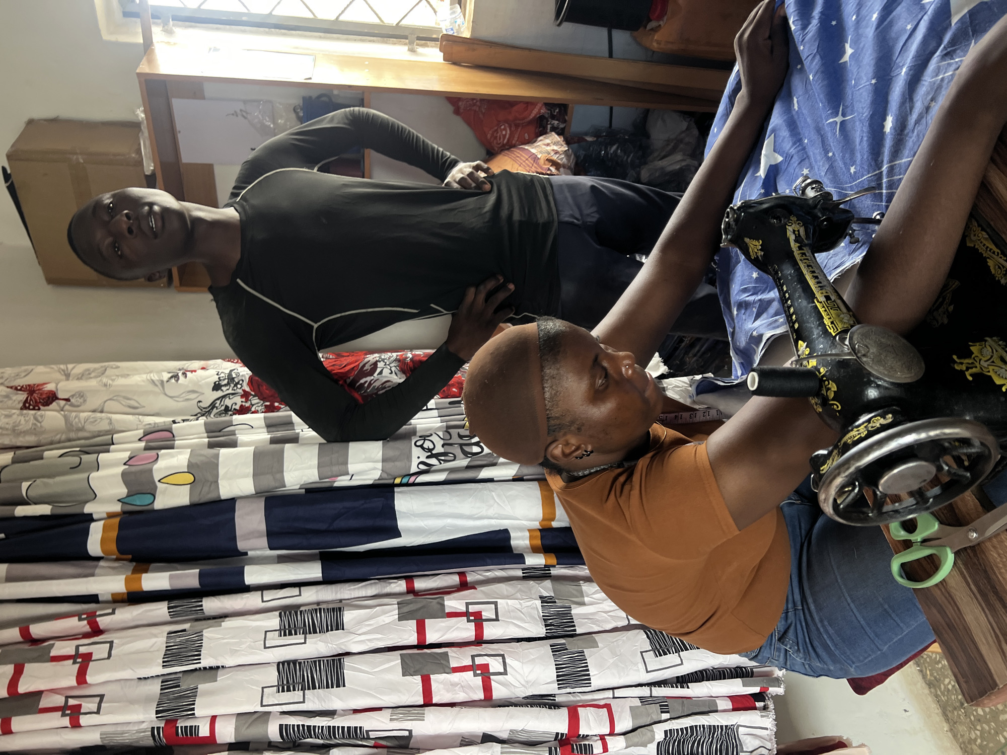 In a room, likely in Jos, Nigeria, a person is seated at a vintage sewing machine, operating it with their hands. They wear a brown t-shirt, blue jeans, and a brown head covering. Next to the sewing machine, a pair of green-handled scissors lies on a wooden surface. Behind them, a younger person stands with hands on their hips, wearing a black long-sleeved shirt and dark pants. The background features a collection of patterned curtains and shelves holding various items, including fabric. The room appears to be a workshop or a stall within a market.