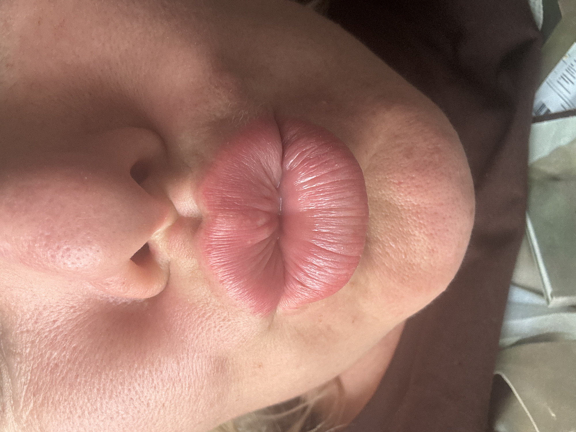 This is a close-up, rotated image focused on the mouth and nose area of a person. The person's lips are pursed, creating deep vertical creases, and are a vibrant pinkish-red color. The skin around the mouth and nose appears to be fair-skinned with visible pores and some very fine, light-colored hairs on the upper lip area. The nostril is also visible. The lighting suggests it is indoors, possibly artificial light given the evenness and slightly warm tone. The background is blurred and dark, with a hint of a textured surface and some faint white text on a light-colored object visible in the upper right corner, but it is illegible. No specific time of day, weather, or explicit emotions are conveyed beyond the action of pursing the lips. The location context of Pozdyshki, Russia, is provided but not visually discernible from the image itself.