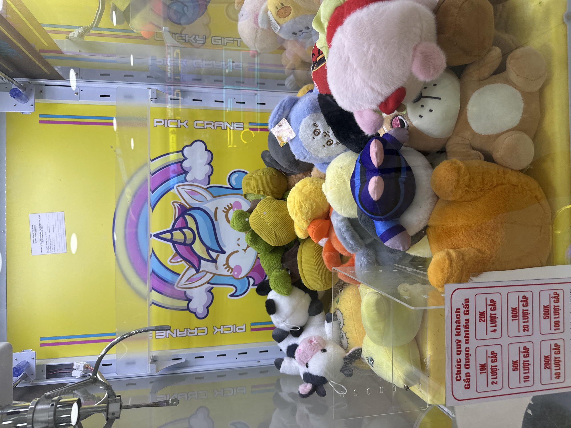 A claw machine filled with stuffed animals is displayed in a store. The machine has a yellow background with a unicorn and rainbow graphic. Several stuffed animals, including a cow, a bear, and a rabbit, are visible inside. A price chart in Vietnamese is attached to the front of the machine. The machine appears to be located in a commercial establishment, possibly a mall or arcade, in Tay Nha Trang Ward, Vietnam.