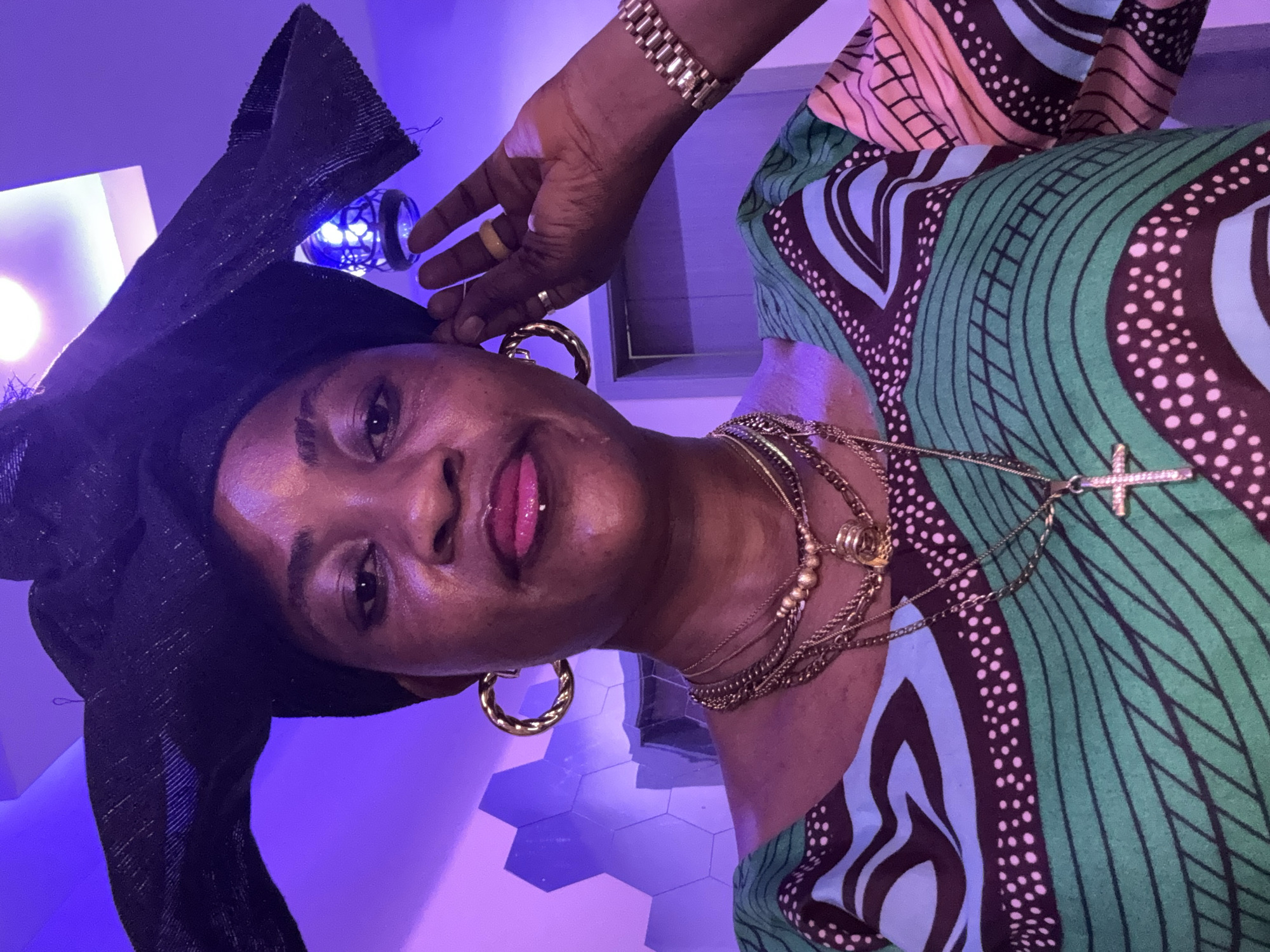 In the city of Ajara, Nigeria, a woman of African descent, adorned in a black headwrap and a vibrantly patterned green, blue, and pink garment, poses for a selfie. She wears multiple gold necklaces, a gold bracelet, and large gold hoop earrings. Her right hand is raised to her ear, while her left hand is extended upwards, touching her forehead. The background is illuminated by a purple light, casting an ambient glow on a textured wall. A decorative lamp hangs from the ceiling.
