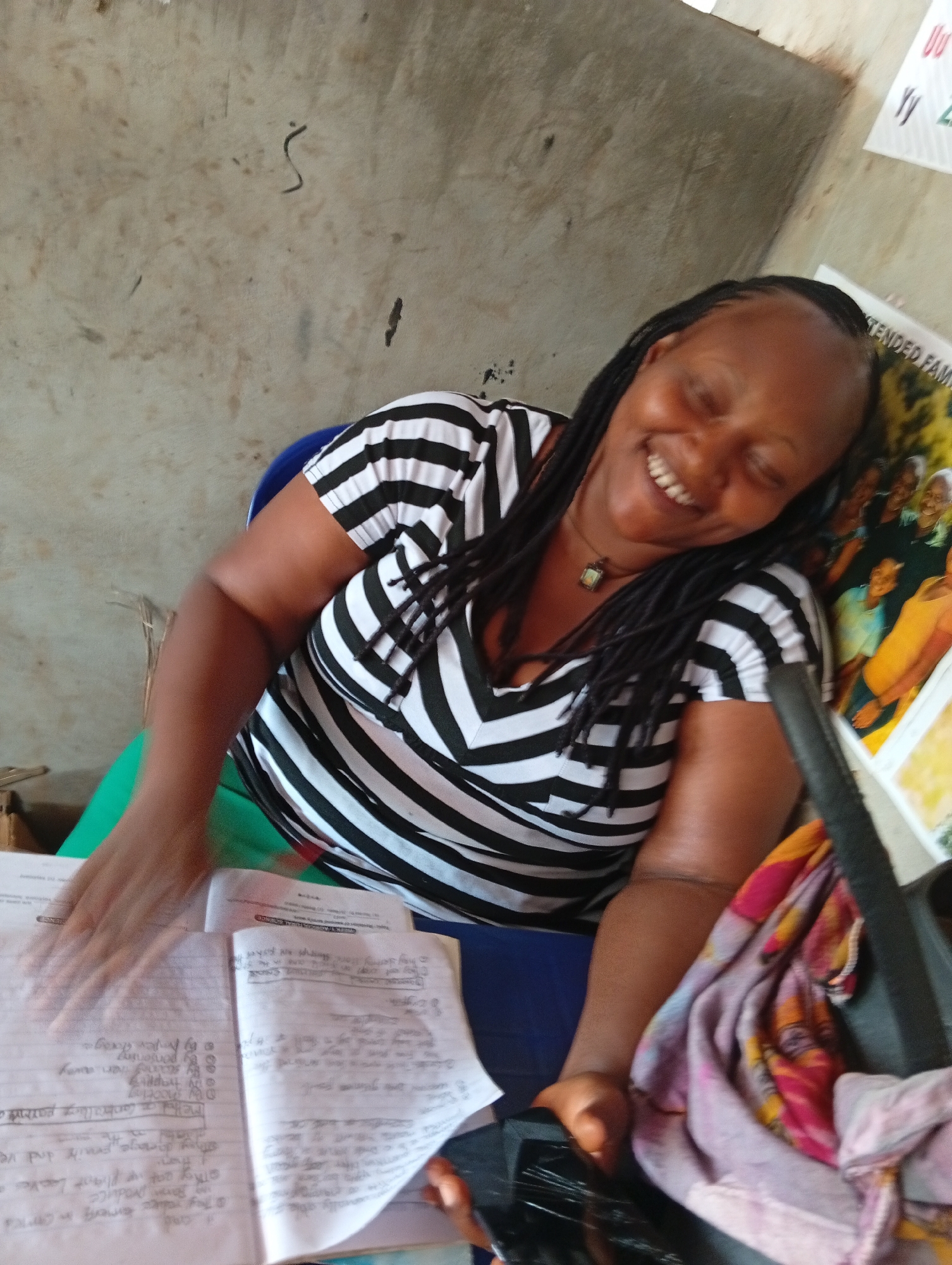 In what appears to be an indoor setting in Ukwu-Abwa, Nigeria, a smiling Black woman with braided hair sits at a desk, wearing a black and white striped shirt. She is seated in a blue and green chair with books and papers on the desk in front of her. A multicolored cloth is draped on the side of the desk. A poster or artwork is visible on the wall behind her, along with some writing.