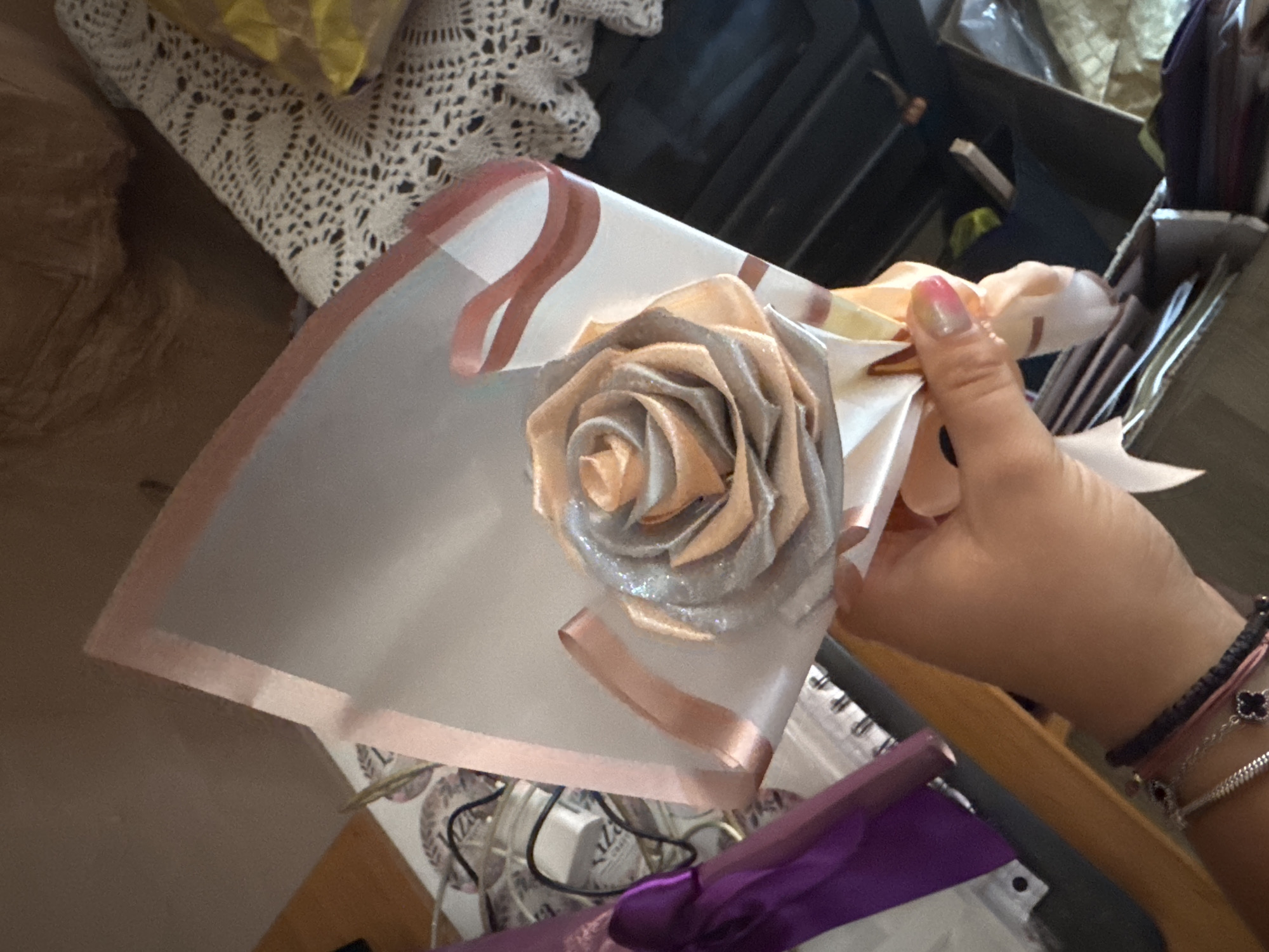 A person's hand is holding a bouquet wrapped in transparent paper, adorned with pink ribbons. The bouquet contains a single, intricately folded rose made of satin fabric, with silver glitter accents. In the background, a white crocheted doily is visible on a surface. To the left, a purple metallic ribbon or fabric is partially visible. The environment suggests an indoor setting, possibly a home or a craft space, with various items in the background including what appears to be storage containers and other crafting materials. The lighting suggests it is daytime. The image does not provide any specific visual information about the city of Sredets, Bulgaria.