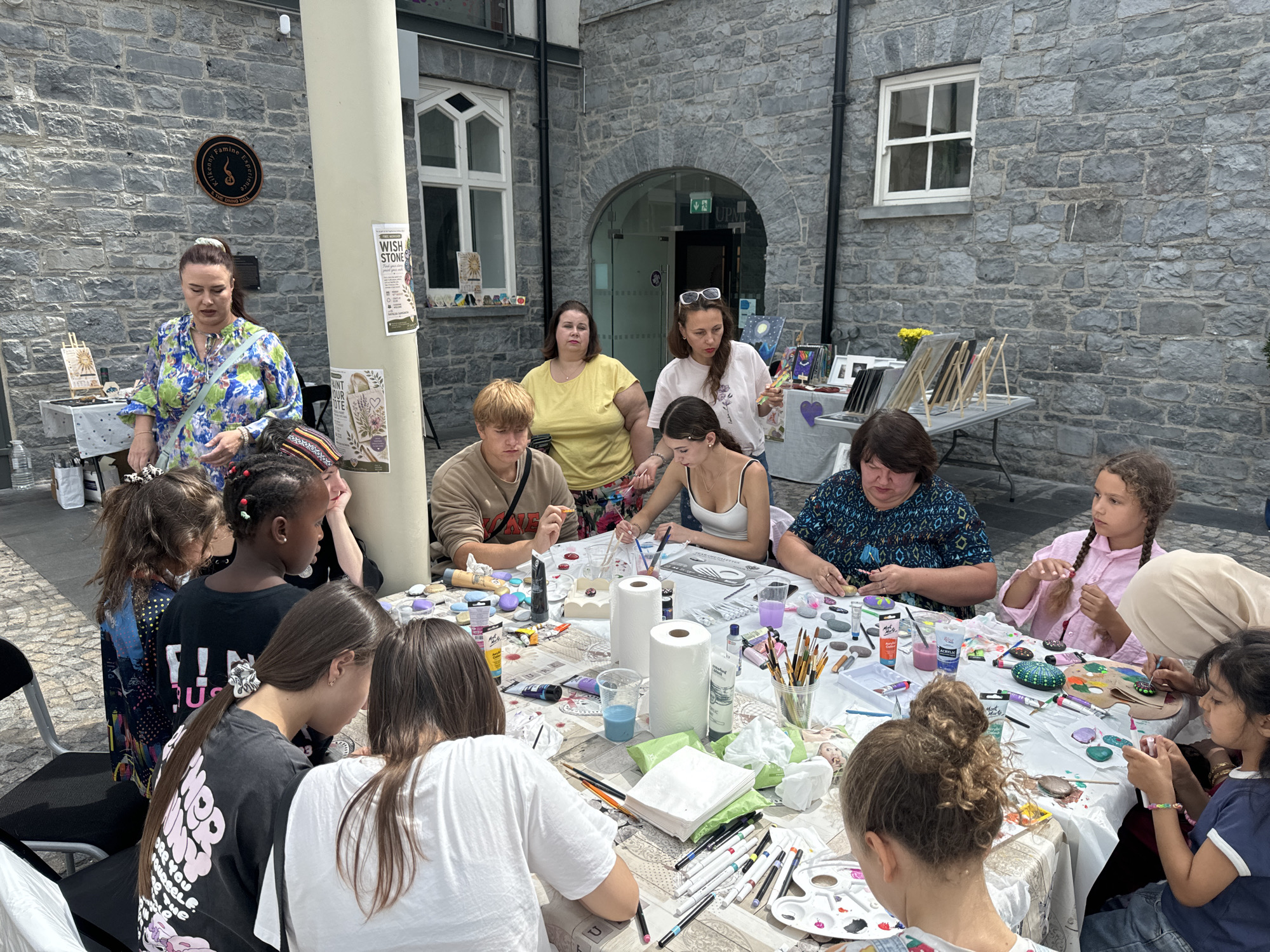 This image captures an outdoor art workshop taking place at the Medieval Mile Museum in Kilkenny, Ireland. The museum's stone facade and arched entrance are visible in the background. A diverse group of individuals, including adults and children, are gathered around a large table covered with art supplies. They are actively engaged in painting, with various paints, brushes, rocks, and paper towels spread across the table. The atmosphere appears to be one of creative collaboration and enjoyment. The workshop is likely part of a community event or educational program organized by the museum.