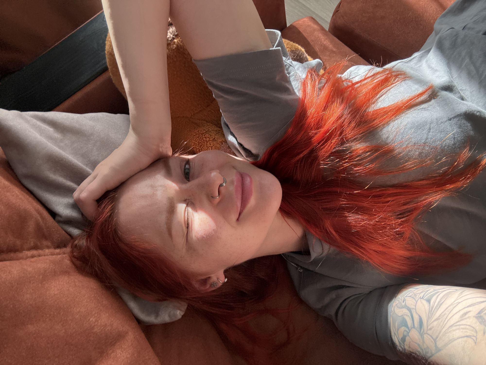 A woman with red hair is lying on a brown couch, her right hand resting on her forehead. She is wearing a grey shirt. She has a nose ring and an earring in her left ear. The couch is made of brown fabric, and there is a black strap visible in the upper left corner. There is a teddy bear behind her head. The woman has a tattoo on her right arm. The image is taken in bright sunlight. The location of the image is not discernible from the photograph and cannot be determined to be Rostov-on-Don, Russia.