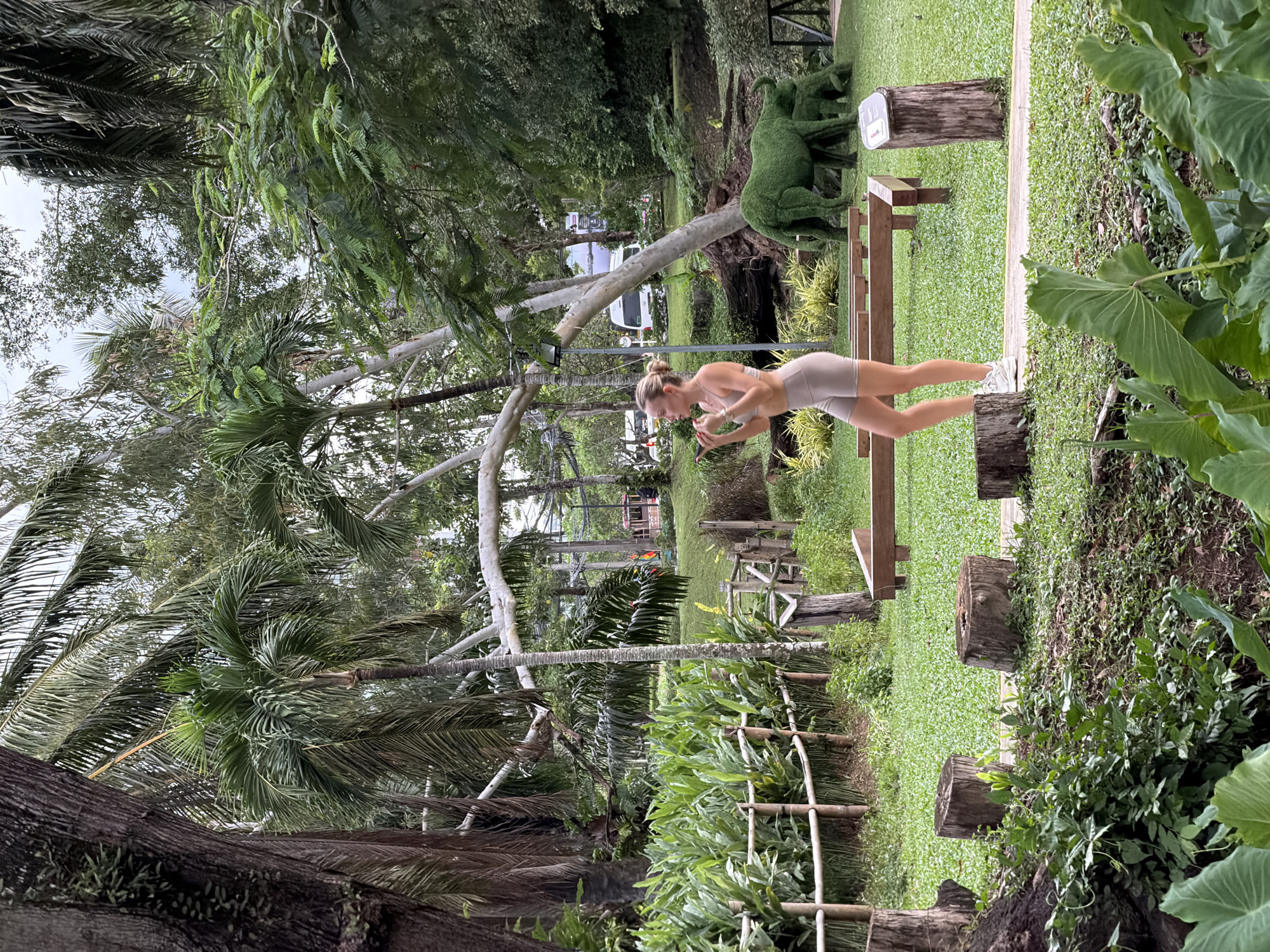 The image depicts a woman in athletic wear standing on a paved path in a park-like setting. She is wearing a light-colored sports bra and matching shorts, and white athletic shoes. Her blonde hair is pulled back. She appears to be holding a phone to her ear and gesturing with her other hand. The background features lush greenery, palm trees, and a variety of other foliage. In the middle ground, there are several tree stumps, a wooden bench, and two topiary sculptures resembling elephants. In the distance, a train-like vehicle with colored cars is visible, along with a white van. The scene suggests an outdoor recreational area, potentially a botanical garden or zoo, in Choeng Thale, Thailand.