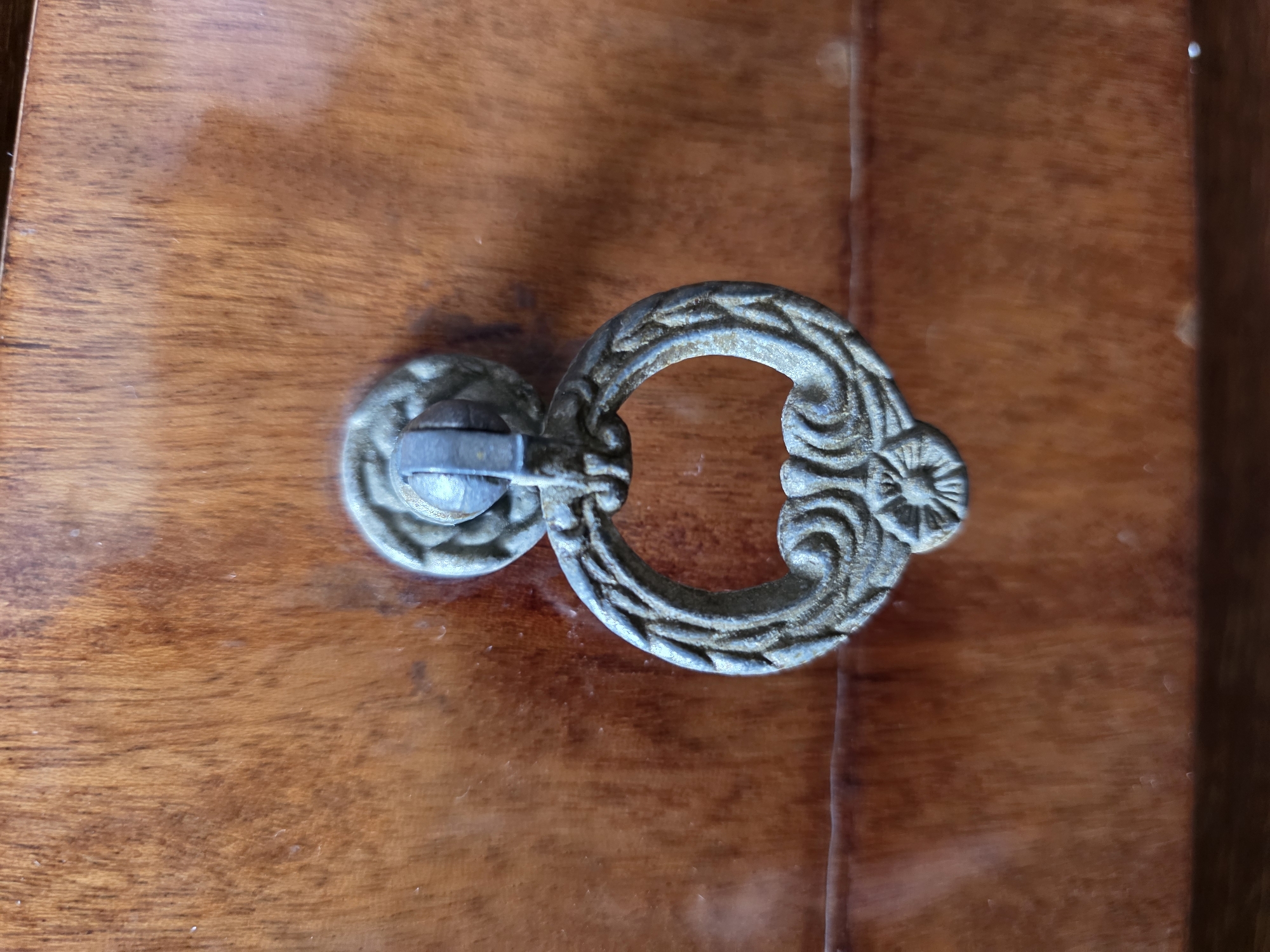 A close-up, eye-level shot shows an ornate, antique drawer pull against a wooden surface. The drawer pull is made of a tarnished, gray metal and features a circular loop with decorative etching and a central flower design. The wooden surface is a warm, medium-brown with visible grain patterns. A horizontal line, possibly the edge of a drawer or shelf, is visible across the lower portion of the image. There are no people or other objects visible. The lighting is soft and natural, creating subtle shadows that highlight the texture of the wood and metal. The scene appears to be indoors, with no indications of an outdoor or city environment.