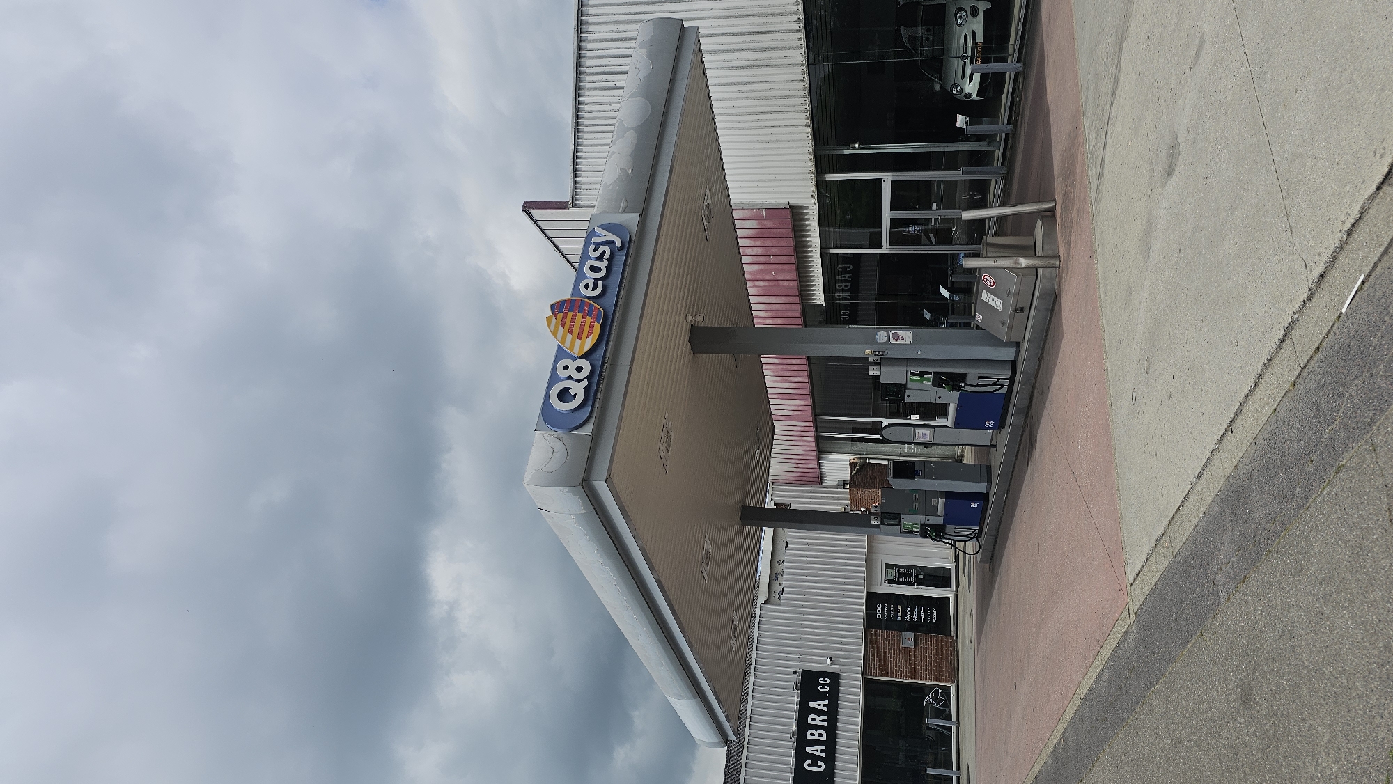 The image shows a Q8 easy gas station on a cloudy day. The gas station is located in Boussu-lez-Walcourt, Belgium. The main structure of the gas station consists of a large canopy supported by pillars. Underneath the canopy are several fuel pumps, some of which are blue and grey. To the left of the gas station, there is a building with a sign that reads "CABRA.cc". Further to the left, there is a storefront with a window displaying what appears to be bicycles. To the right of the gas station, through a large glass window, a white car is visible, with the word "OCASION" on its rear. The sky is filled with dark, heavy clouds. The ground in front of the gas station is paved with asphalt and a reddish-pink surface.