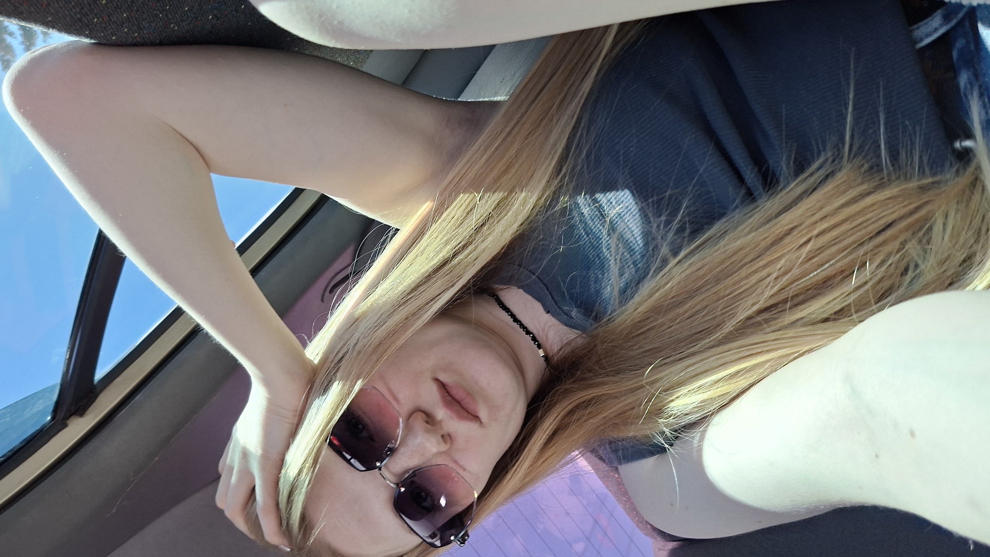 A blonde-haired person wearing sunglasses and a dark shirt is inside a vehicle, possibly a car, with a visible window and a view of the sky. The location in Прикамский, Russia, is not discernible from the image.