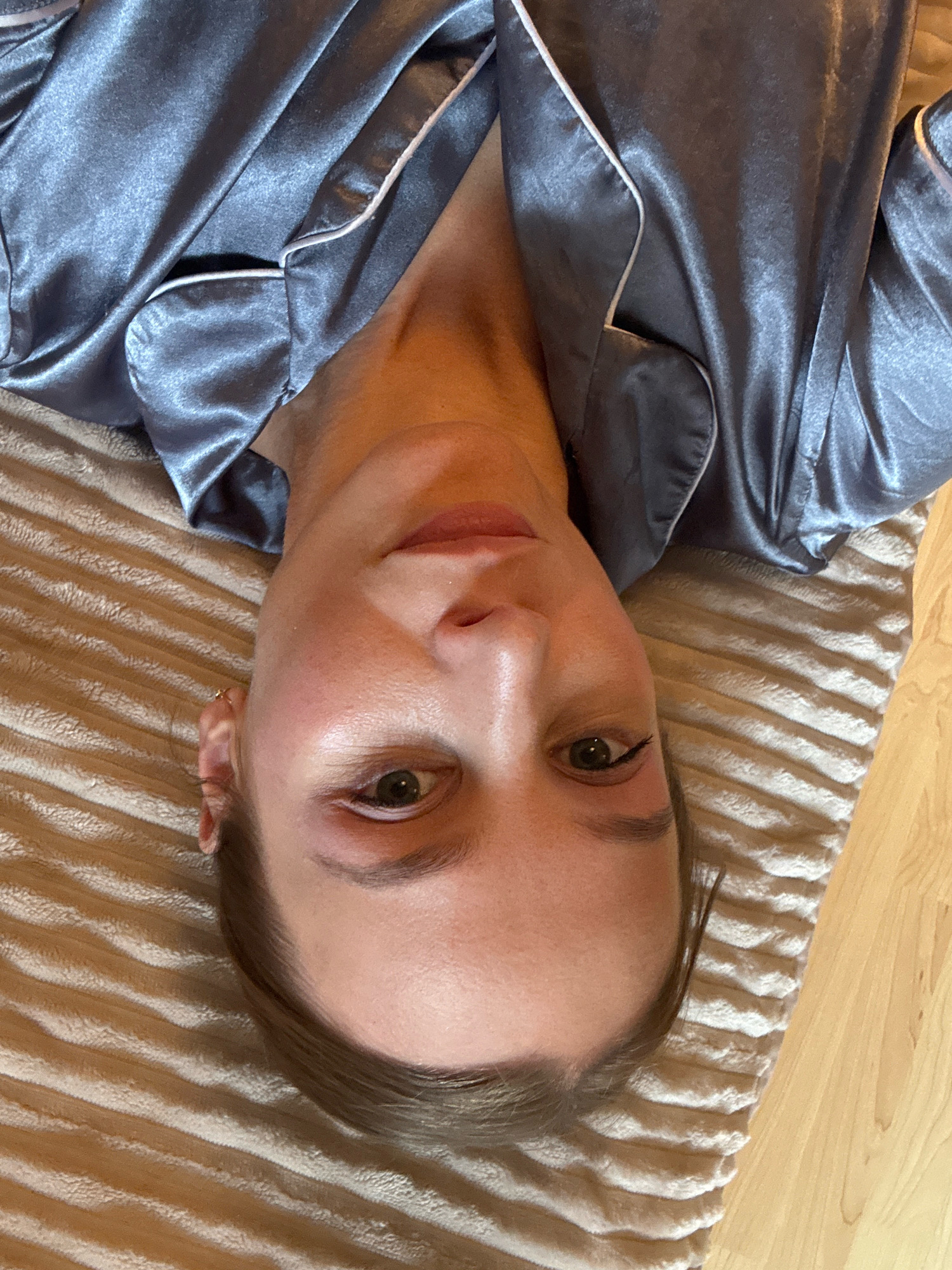 A person is lying down on a striped, tan-colored textured blanket. The person is wearing a satin pajama top that is grey with white piping. The top is unbuttoned, revealing the person's neck and chest. The person's face is visible, and they appear to be looking at the camera. The image is taken from a slightly elevated angle, looking down at the person. The background is a light brown wooden surface. There is no information about the specific location in the city of Überherrn, Germany.