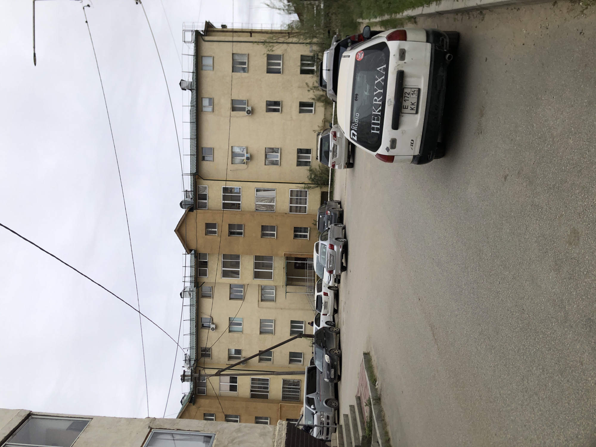 The image shows a residential building with a light yellow facade and a green roof. The building has multiple stories and many windows. In front of the building, there is a parking lot with several cars. A white station wagon is parked in the foreground, with the text "HEKRYXA" visible on its rear window. Other vehicles include a silver van, a dark-colored SUV, and several sedans. The ground is paved with asphalt. The sky is overcast. Based on the presence of the license plate format "E 172 KK 14", this scene is likely located in Yakutsk, Russia, as this format is used in the Sakha Republic.