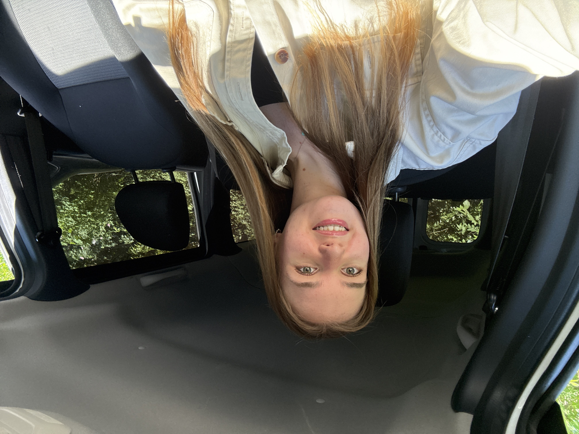 A woman is upside down in the back of a car. Her long, light brown hair hangs down towards the ceiling of the vehicle. She has fair skin, green eyes, and is smiling. She wears a light-colored button-down shirt. The back seats of the car are black and gray. The car windows are open, revealing green foliage and trees outside. The interior of the car is light gray. There is no indication this photo was taken in Perm, Russia.