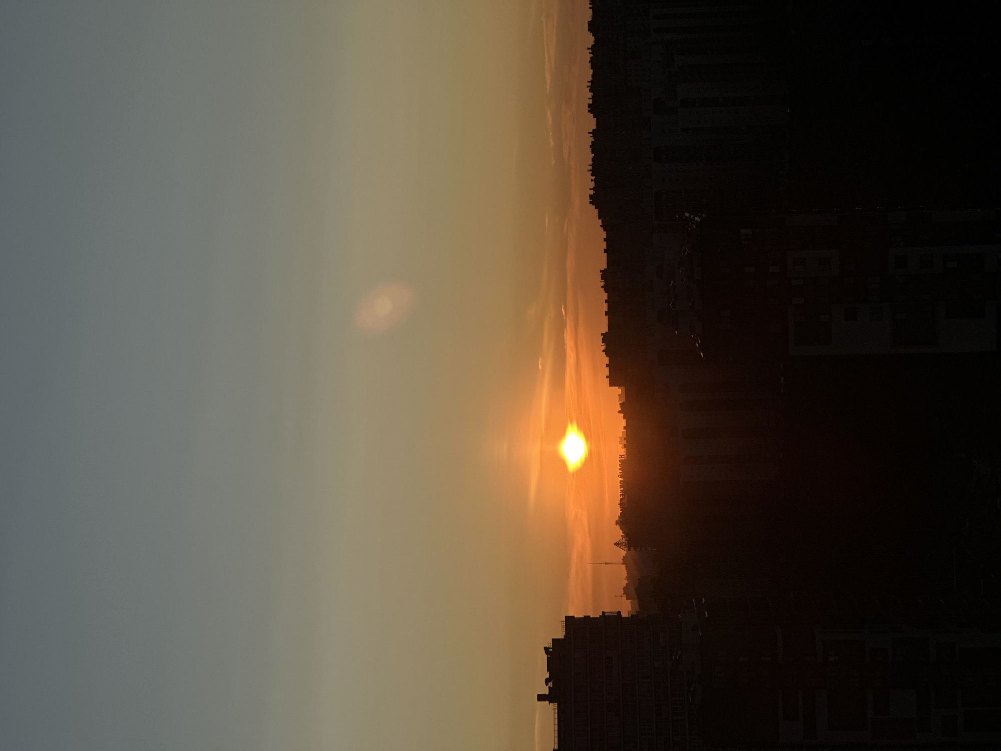 A vertical, eye-level, medium shot captures a city skyline at sunset. The sky transitions from a pale blue at the top to a gradient of orange and yellow towards the horizon. A bright, setting sun, appearing as a white-hot orb, is positioned in the center-left of the image, casting a warm glow. A faint, oval lens flare is visible in the upper right quadrant of the sky. Silhouetted against the vibrant sunset are the dark forms of various buildings, creating a stark contrast between the illuminated sky and the shadowed cityscape. The buildings range from tall, modern structures on the left to lower-profile, densely packed rooftops on the right. In the mid-ground, faint outlines of other structures and possibly trees are visible, appearing as dark shapes against the sunset's light. The overall scene conveys a sense of urban tranquility as day transitions into night.