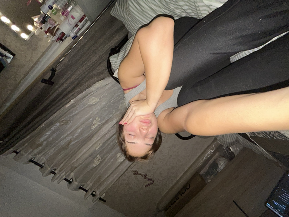 A young woman lies on a striped bed wearing a gray tank top and dark leggings. She covers her face with her hand, displaying red nail polish. Behind her, sheer curtains hang beside a wall with the word "love" displayed. In the upper left, a vanity with lights is visible. The image is oriented at an angle, with the woman positioned diagonally.