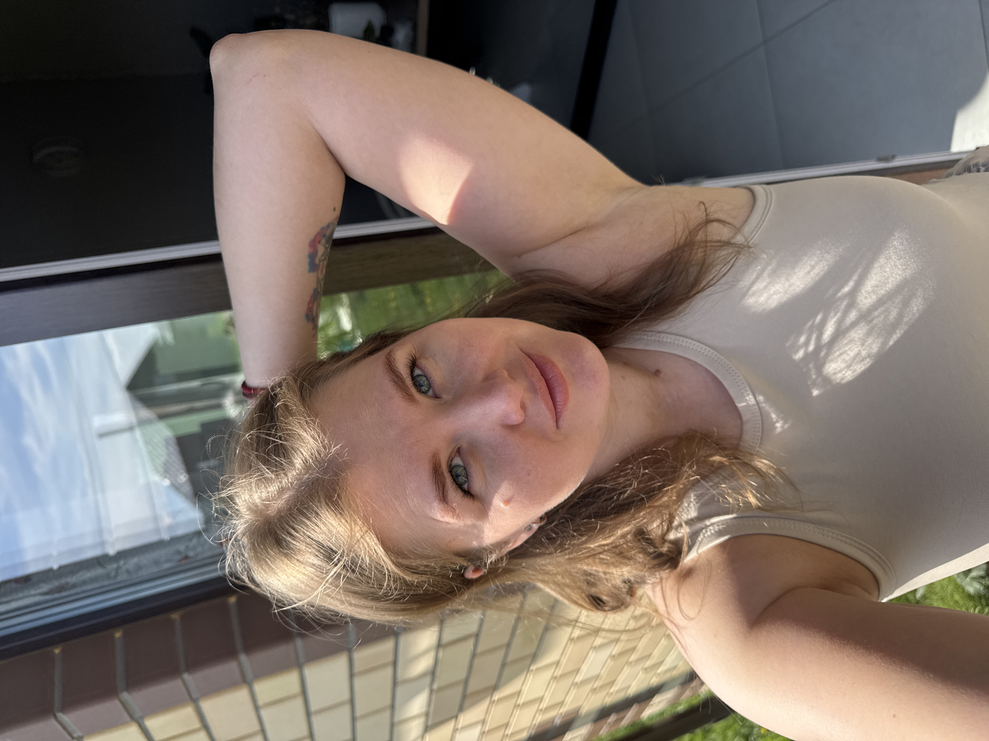 A young woman with fair skin and light brown hair is pictured in a close-up selfie. She has her arm raised and bent behind her head. She is wearing a light-colored tank top. Her eyes are light blue. A colorful tattoo is visible on her upper arm. The background includes a window and a view of what appears to be an outdoor area with greenery and brickwork. The photo might be taken in the city of Ферма, Russia, judging by the prompt.
