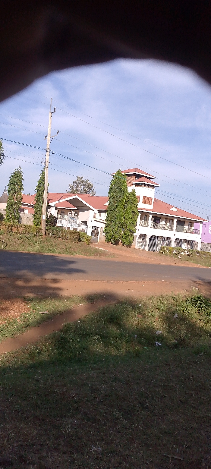This outdoor photograph captures a multi-story building situated alongside a paved road, typical of a semi-urban or rural setting in Bondo, Kenya. The dominant subject is a large, white building featuring multiple balconies with decorative white railings and a prominent red tiled roof. A distinctive tower-like structure with several levels and smaller windows rises from one section of the roof. Tall, slender evergreen trees, possibly cypresses, stand in front of and around the building, providing natural landscaping.
In the foreground, a paved road stretches across the mid-section of the image, bordered by dirt shoulders and grassy verges. The grass appears somewhat dry and contains small patches of weeds and scattered litter. A sturdy wooden utility pole, strung with overhead wires, stands prominently on the left side of the road, extending towards the building.
The sky is bright and clear with scattered light clouds, indicating daytime conditions, likely morning or late afternoon. The overall atmosphere is peaceful. On the property boundary of the building, near the gate, a blue sign with white text is partially visible. The top line clearly reads "BOND O", while the second line is less distinct but appears to begin with 'H', possibly spelling out "HOSPITAL" or "HOTEL", suggesting the building's function. A dark, blurry shape, likely a finger or thumb, partially obstructs the upper left corner of the image.