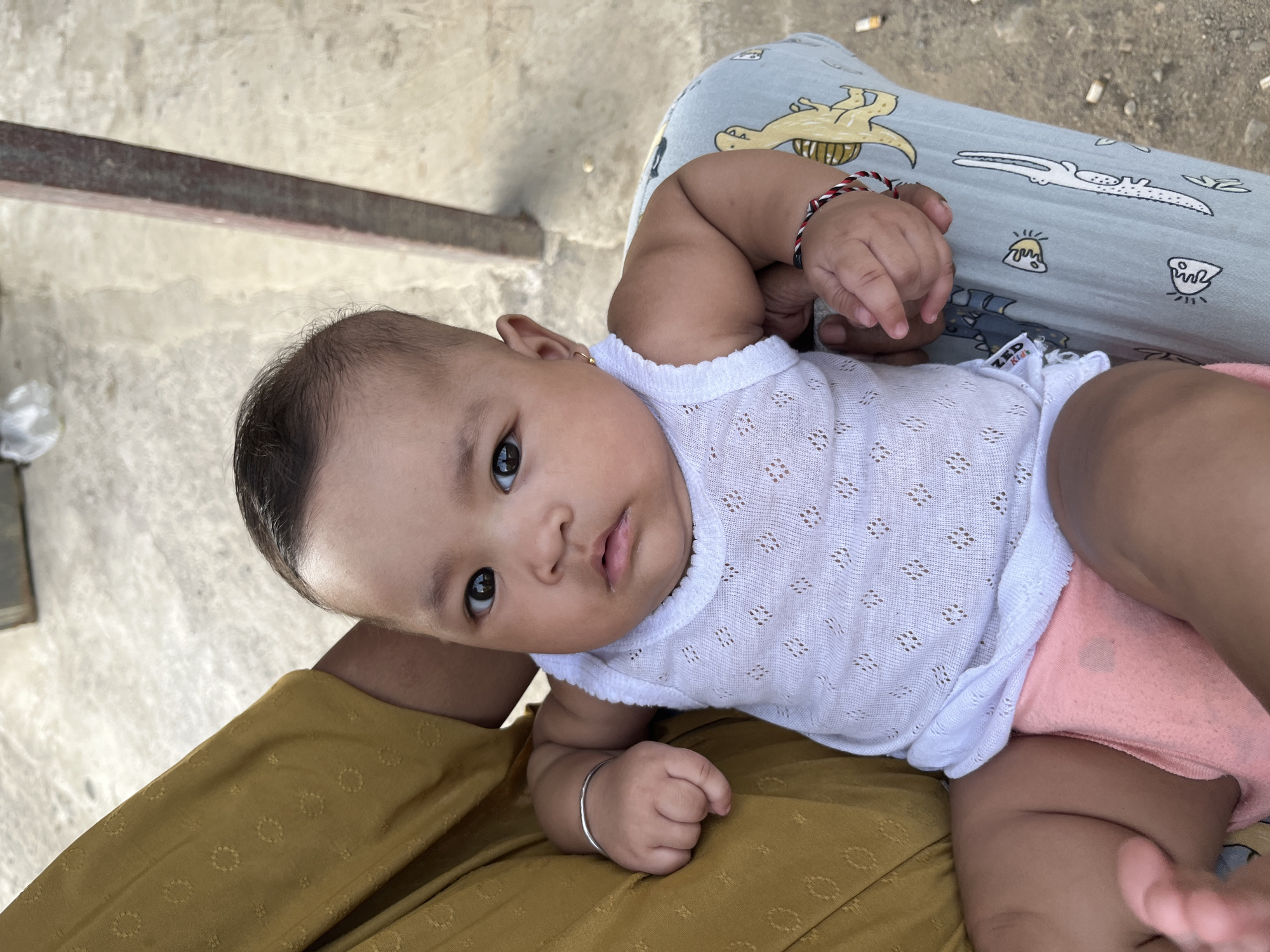 An overhead medium shot captures a dark-skinned baby sitting on someone's lap. The baby is wearing a white tank top with a diamond pattern and pink shorts. The baby's left arm is resting on a yellow patterned fabric, and a silver bracelet is visible on its wrist. The baby's right arm is also resting on a light blue fabric with cartoon dinosaur prints, and a red, white, and black string bracelet is worn on its wrist. The baby's facial expression is neutral, with dark eyes and dark hair. The background is blurry, showing a light-colored concrete surface and a metal pole. This image was taken outdoors in the city of Kruengmate, Indonesia.